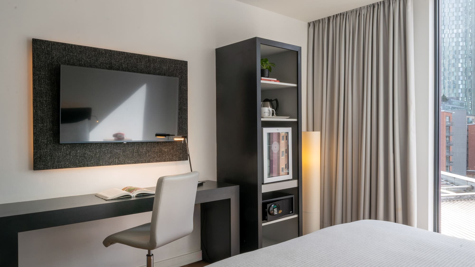 Modern hotel room with desk, TV, mini-fridge, safe, and city view.
