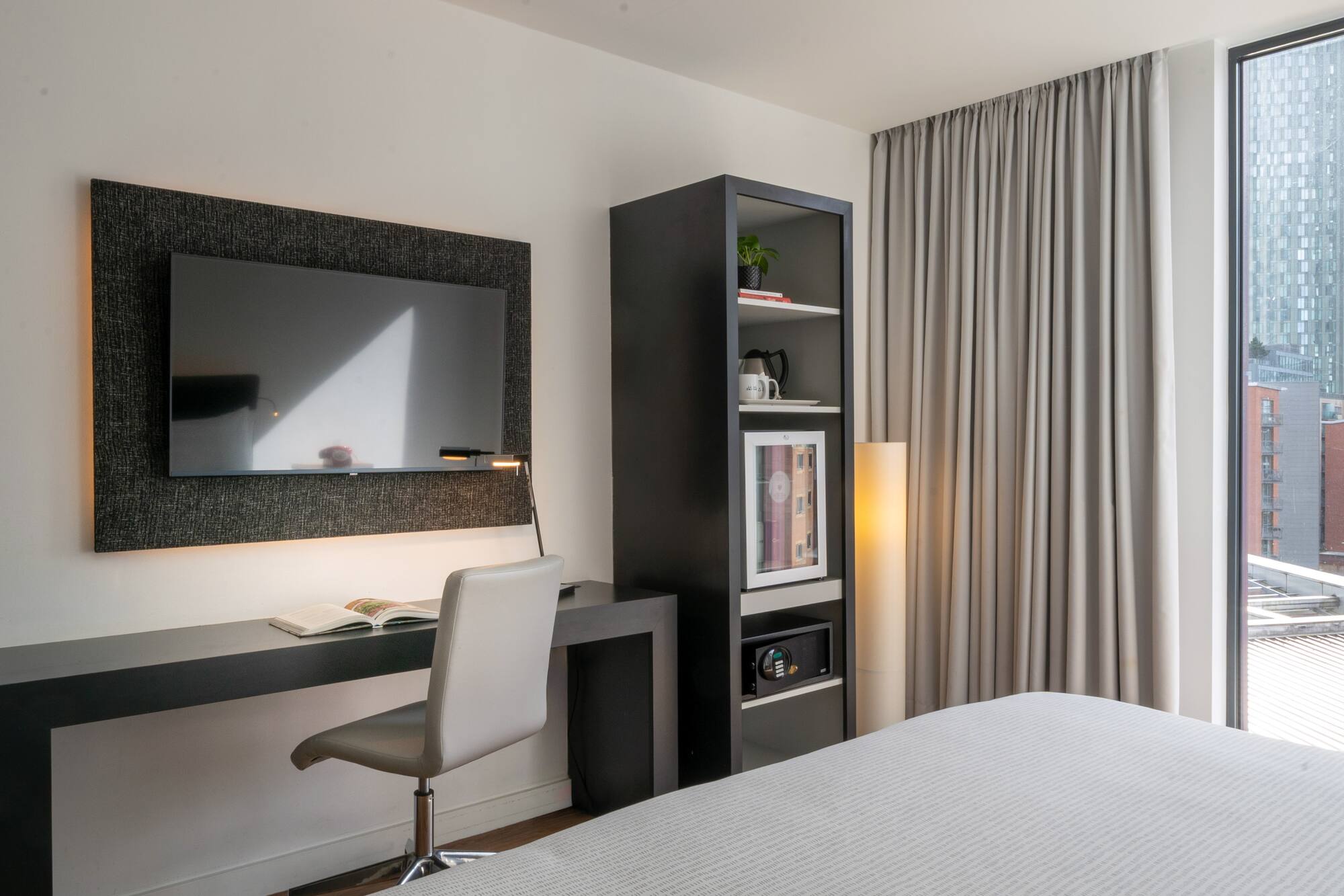 Modern hotel room with desk, TV, mini-fridge, safe, and city view.