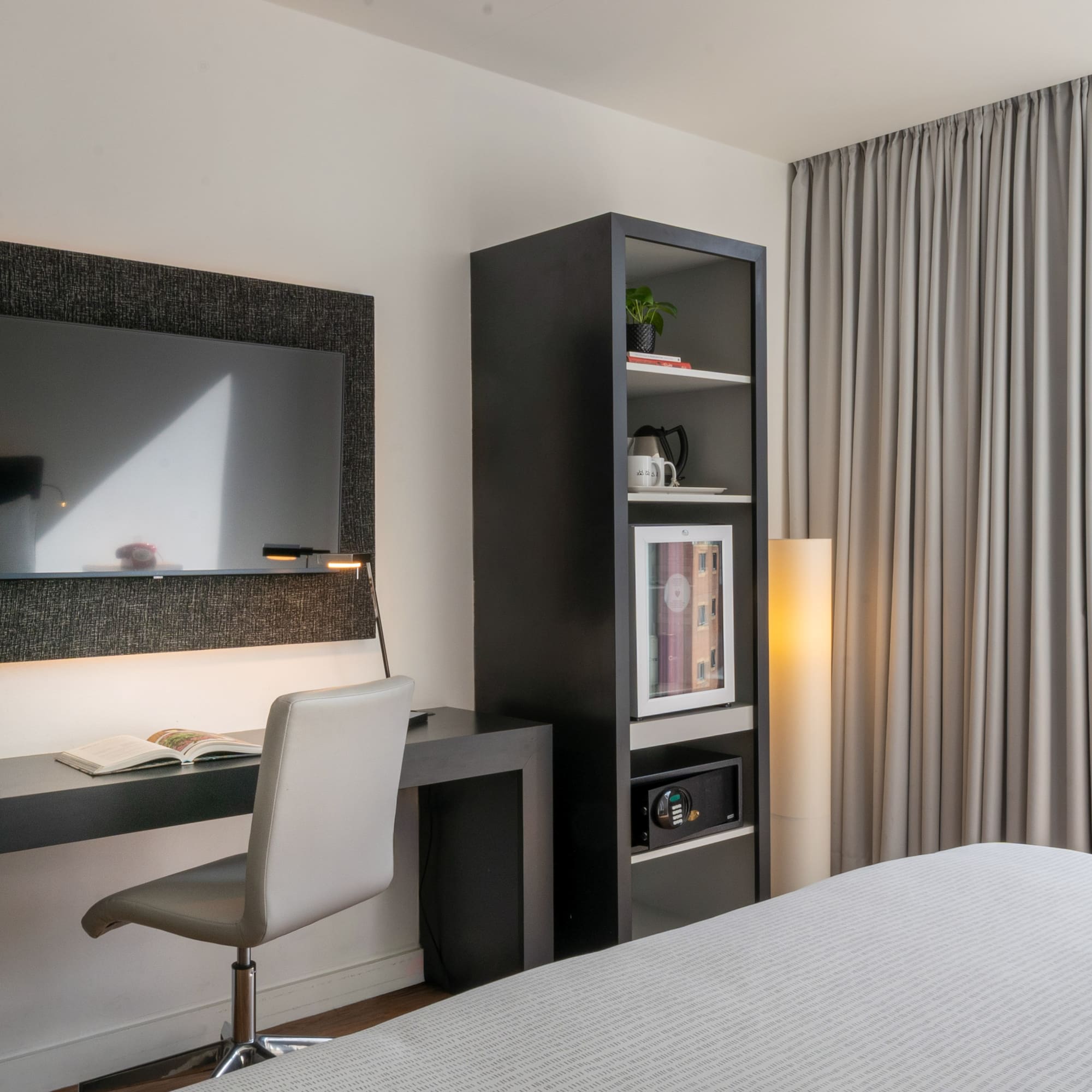 Modern hotel room with desk, TV, mini-fridge, safe, and city view.
