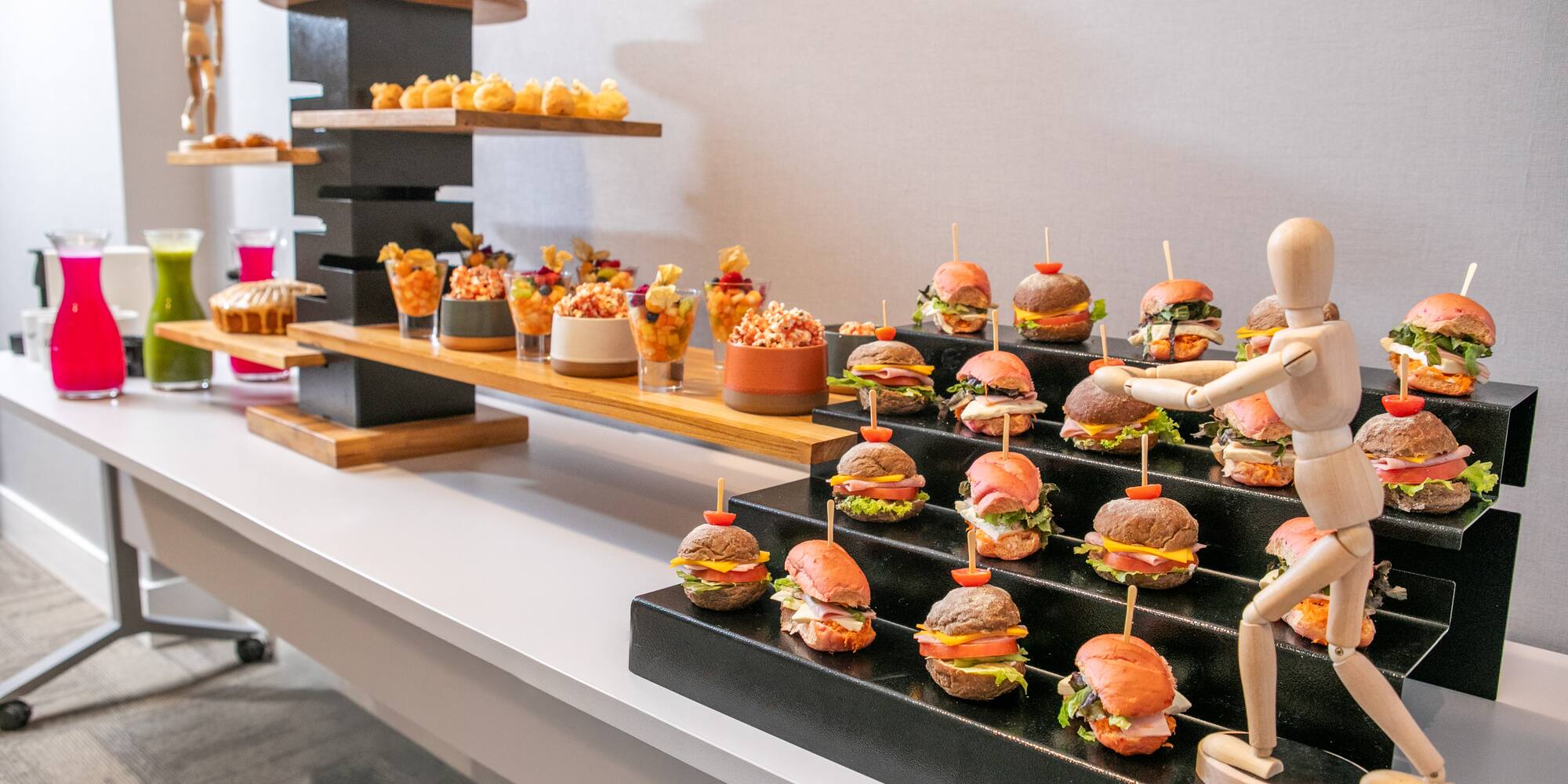 Appetizer buffet with mini burgers, fruit cups, pastries, and colorful juices.