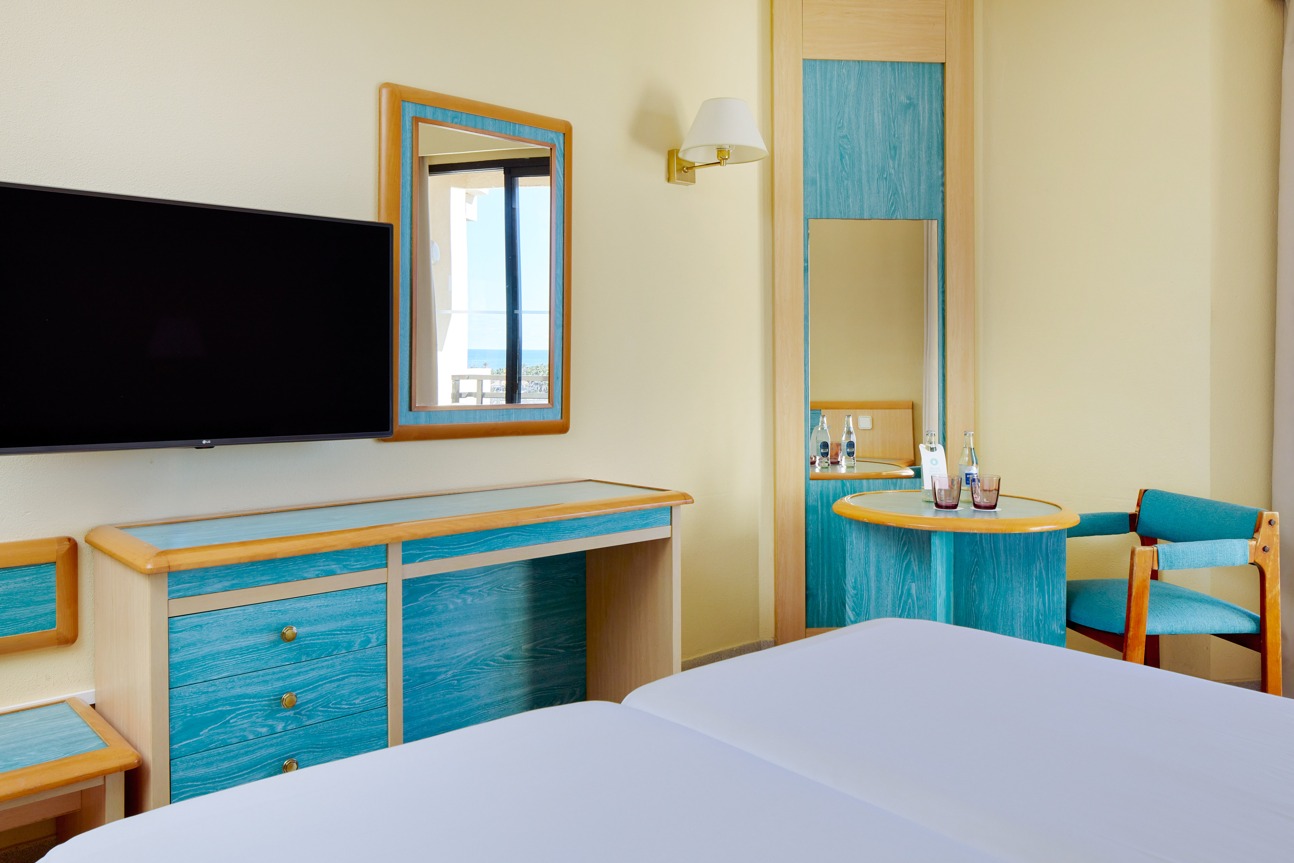 Hotel room with twin beds, TV, teal furniture, and mirror showing sea view.