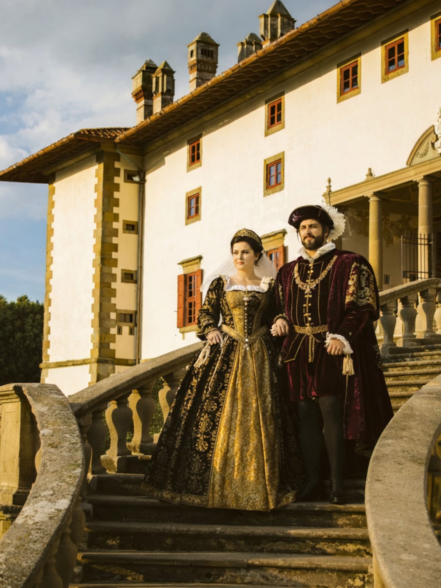Couple in elaborate Renaissance costumes descending grand villa stairs.