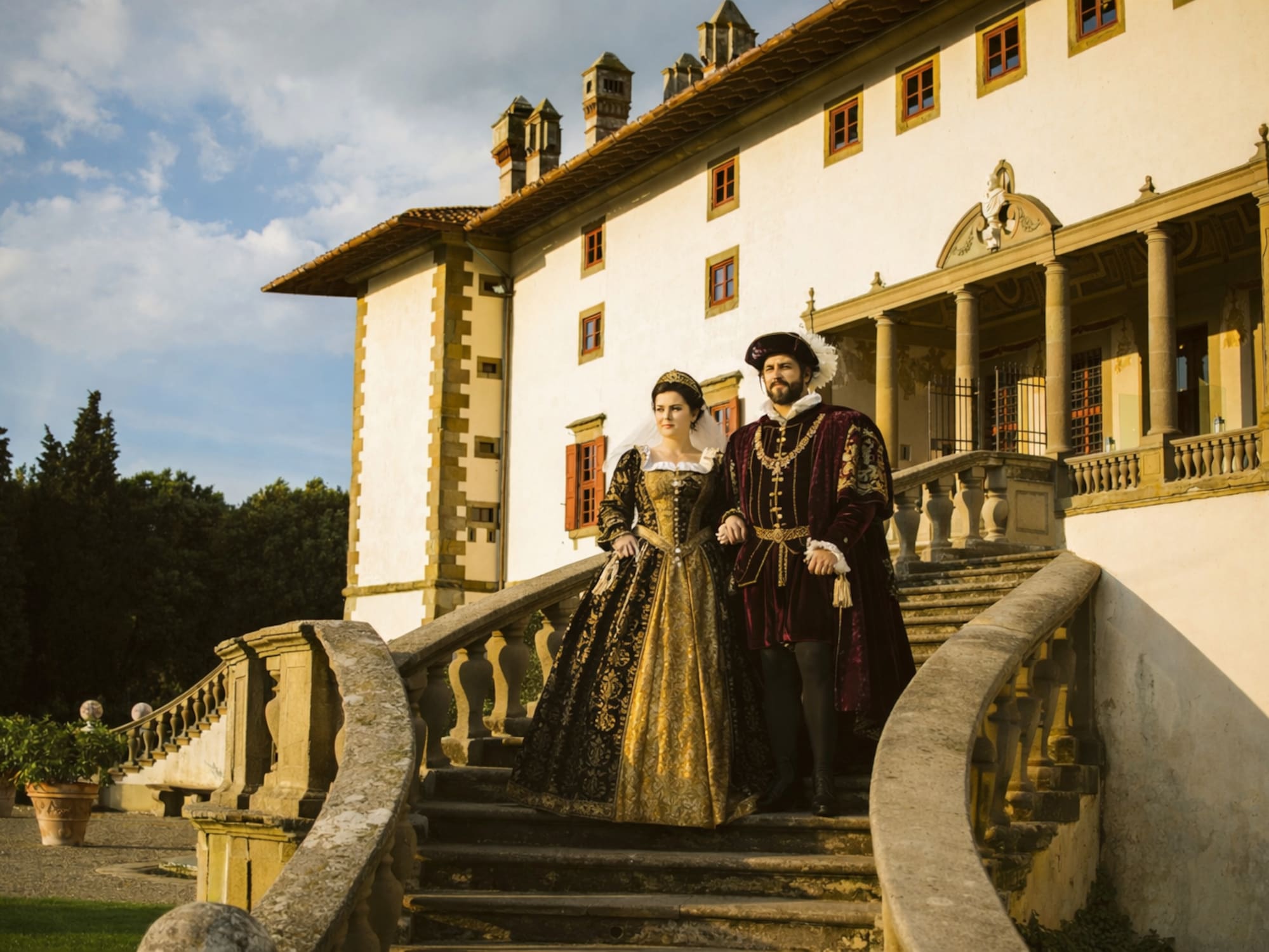 Couple in elaborate Renaissance costumes descending grand villa stairs.