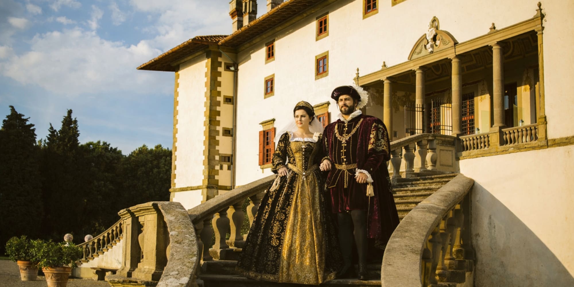 Couple in elaborate Renaissance costumes descending grand villa stairs.