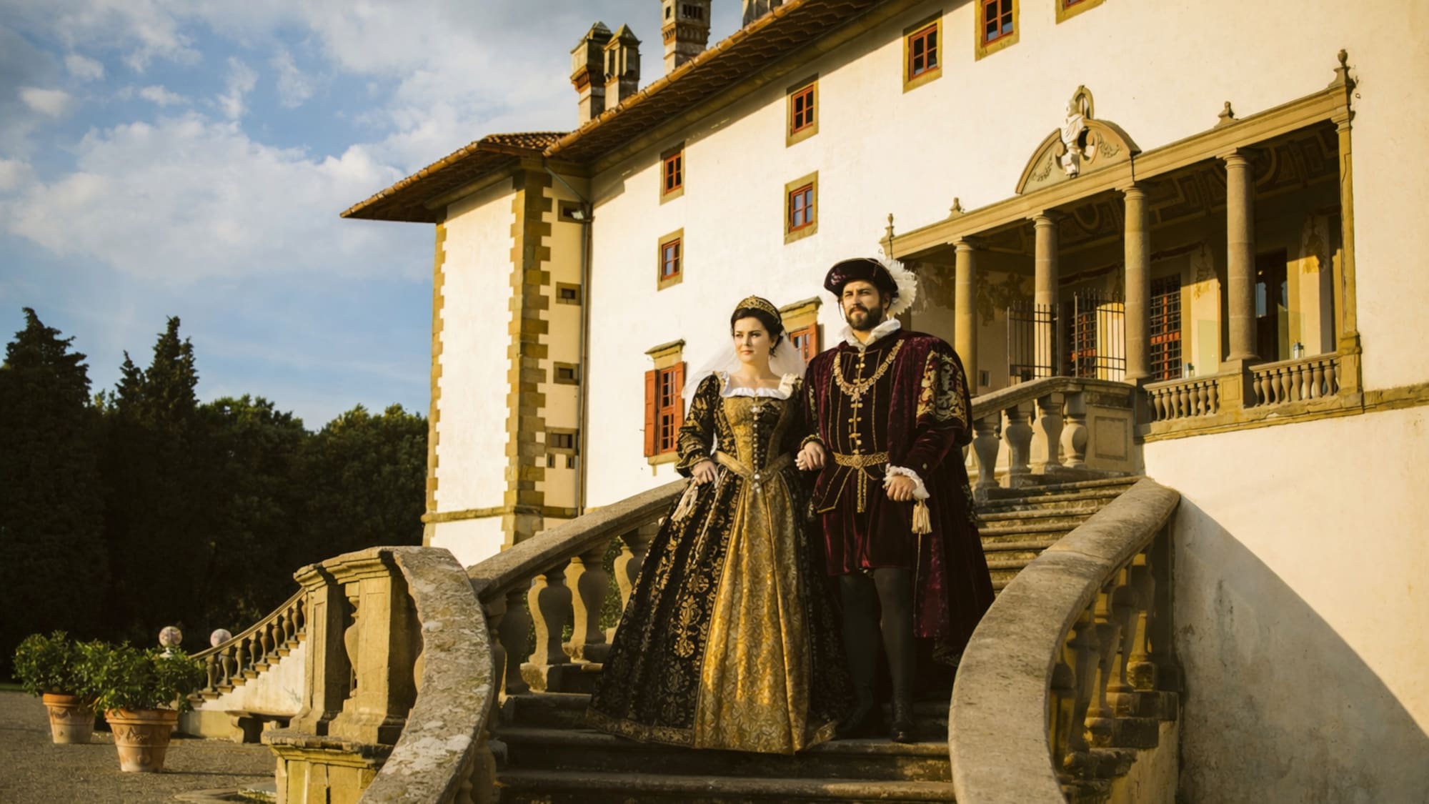 Renaissance couple in period costumes on grand stone stairs of a historic villa.