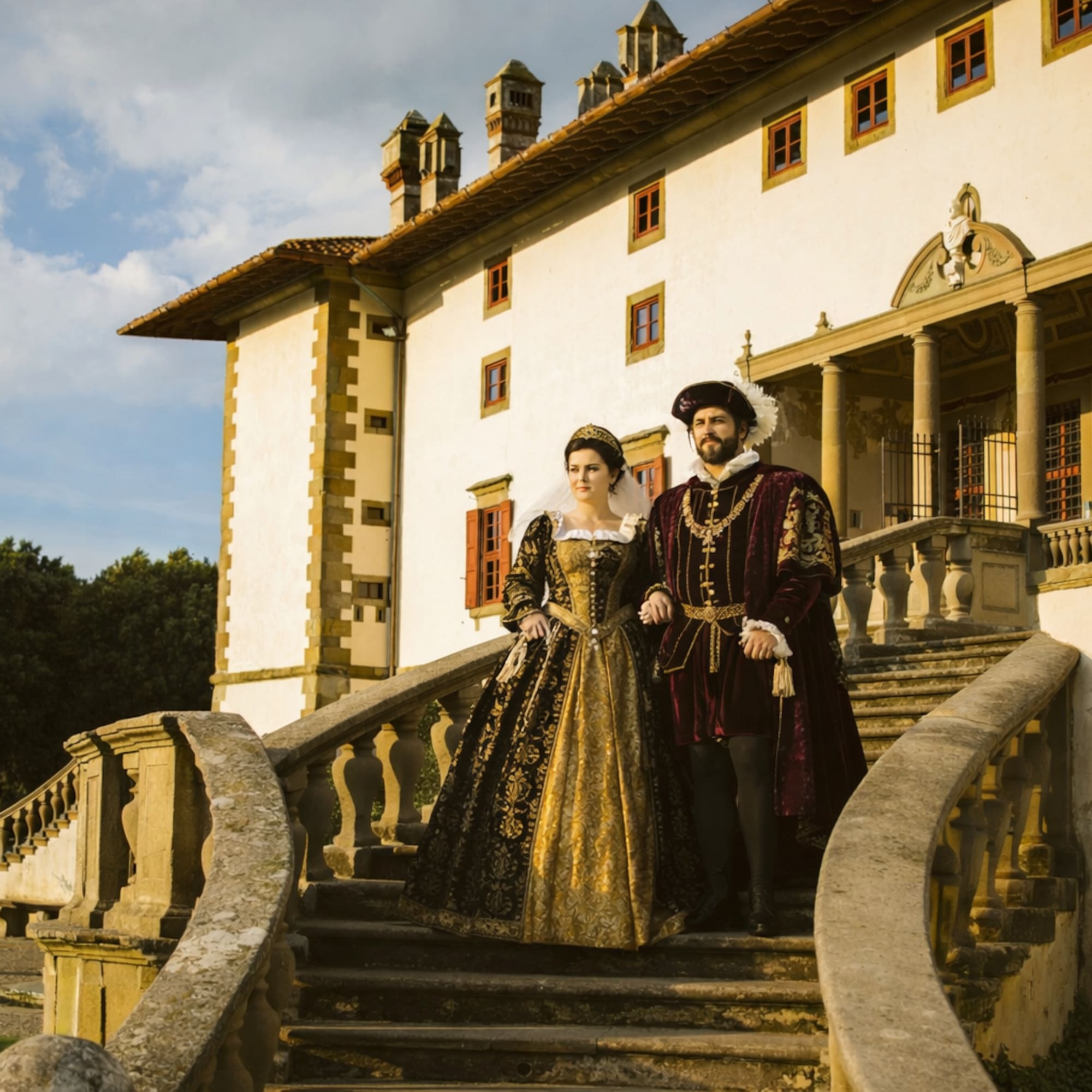 Couple in elaborate Renaissance costumes descending grand villa stairs.