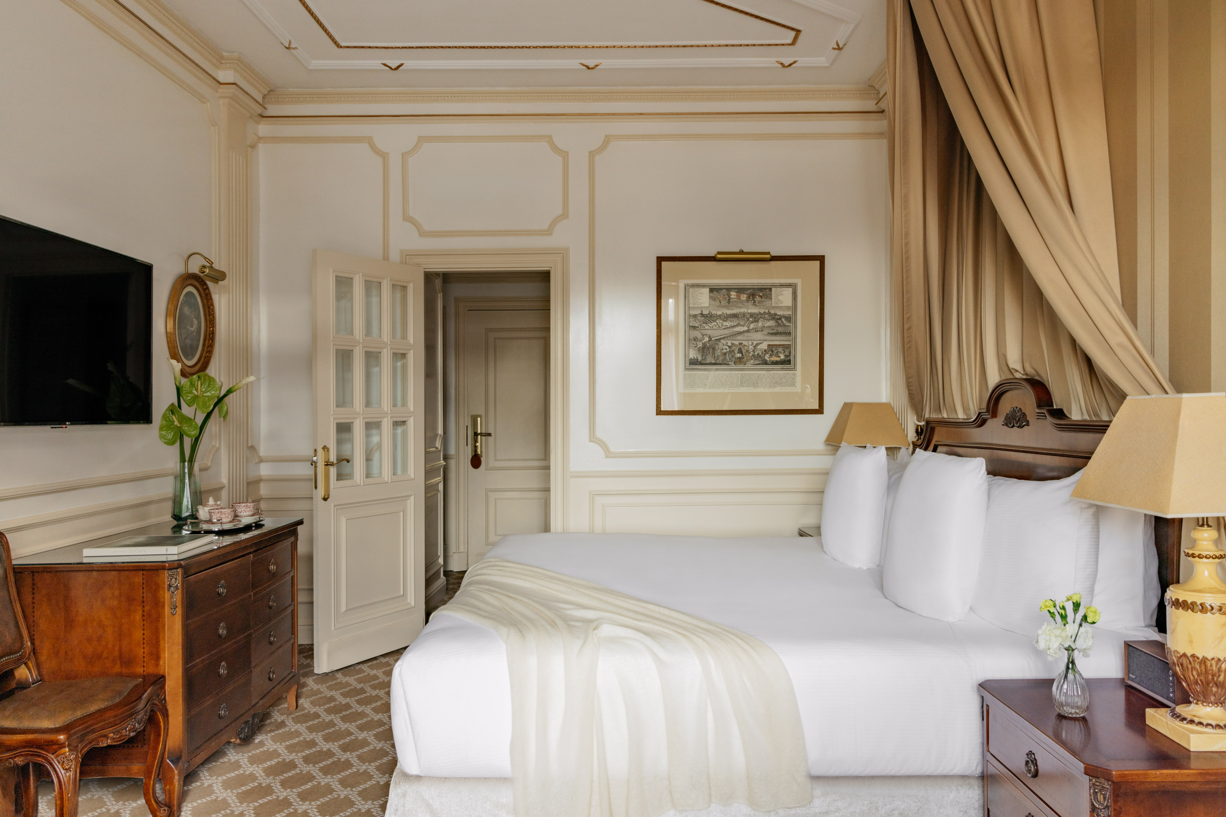 Elegant hotel bedroom with white king bed, ornate wood furniture, and gold accents.