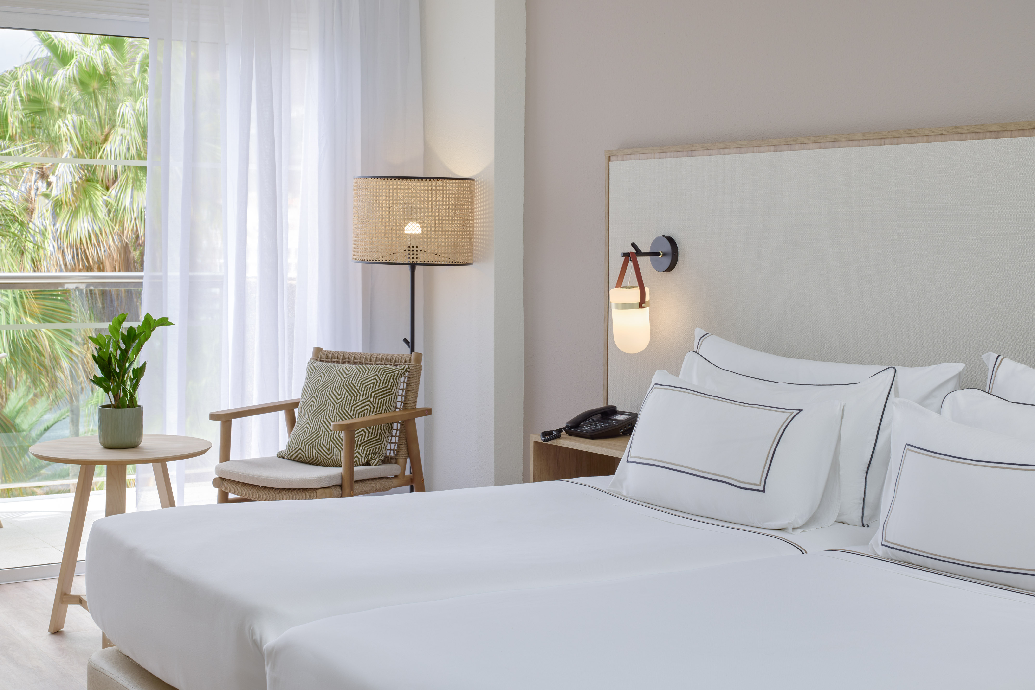 Modern hotel bedroom with white beds, rattan lamp, wooden chair, and tropical view.