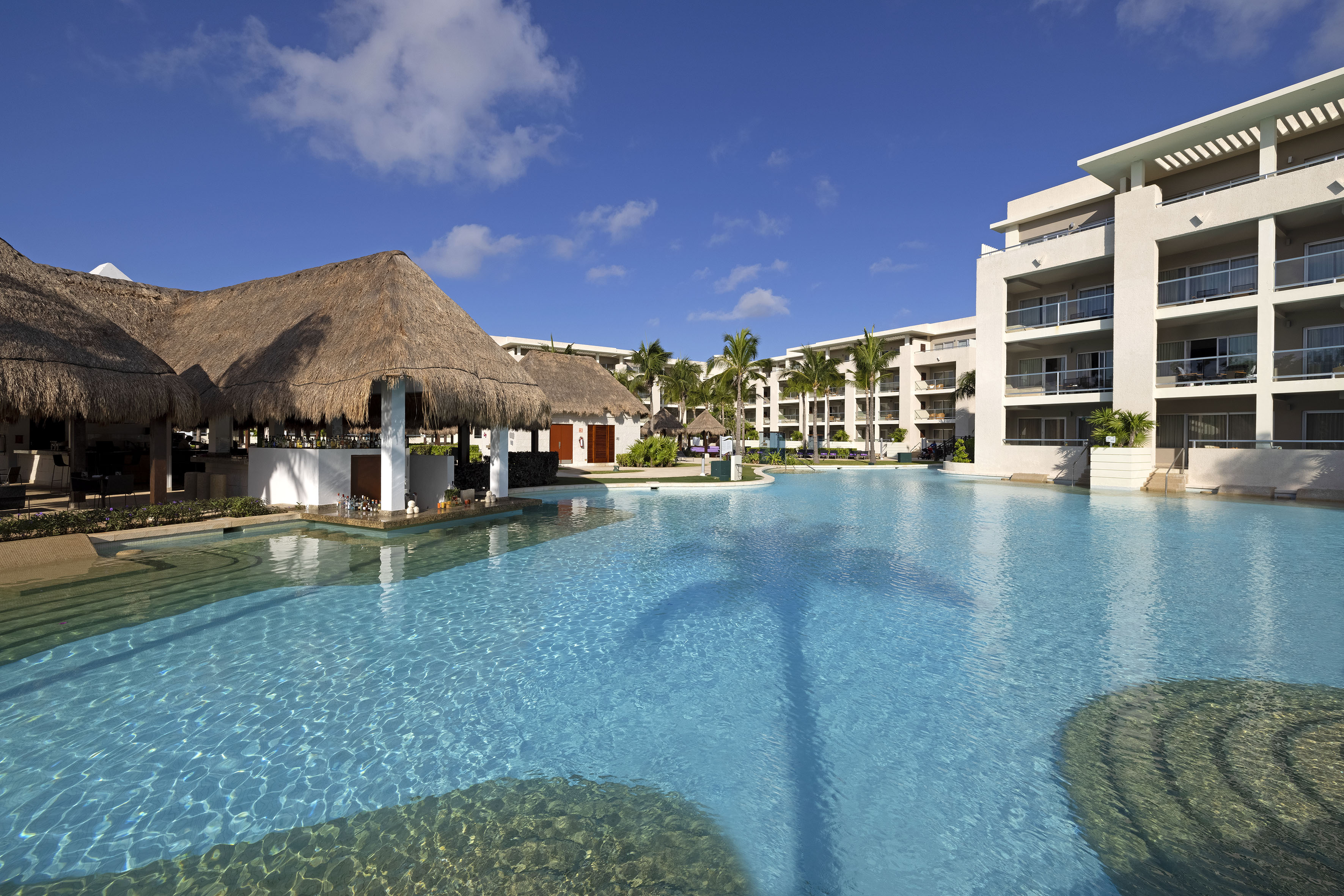 Tropical resort pool with swim-up bar, palm trees, and modern hotel buildings.