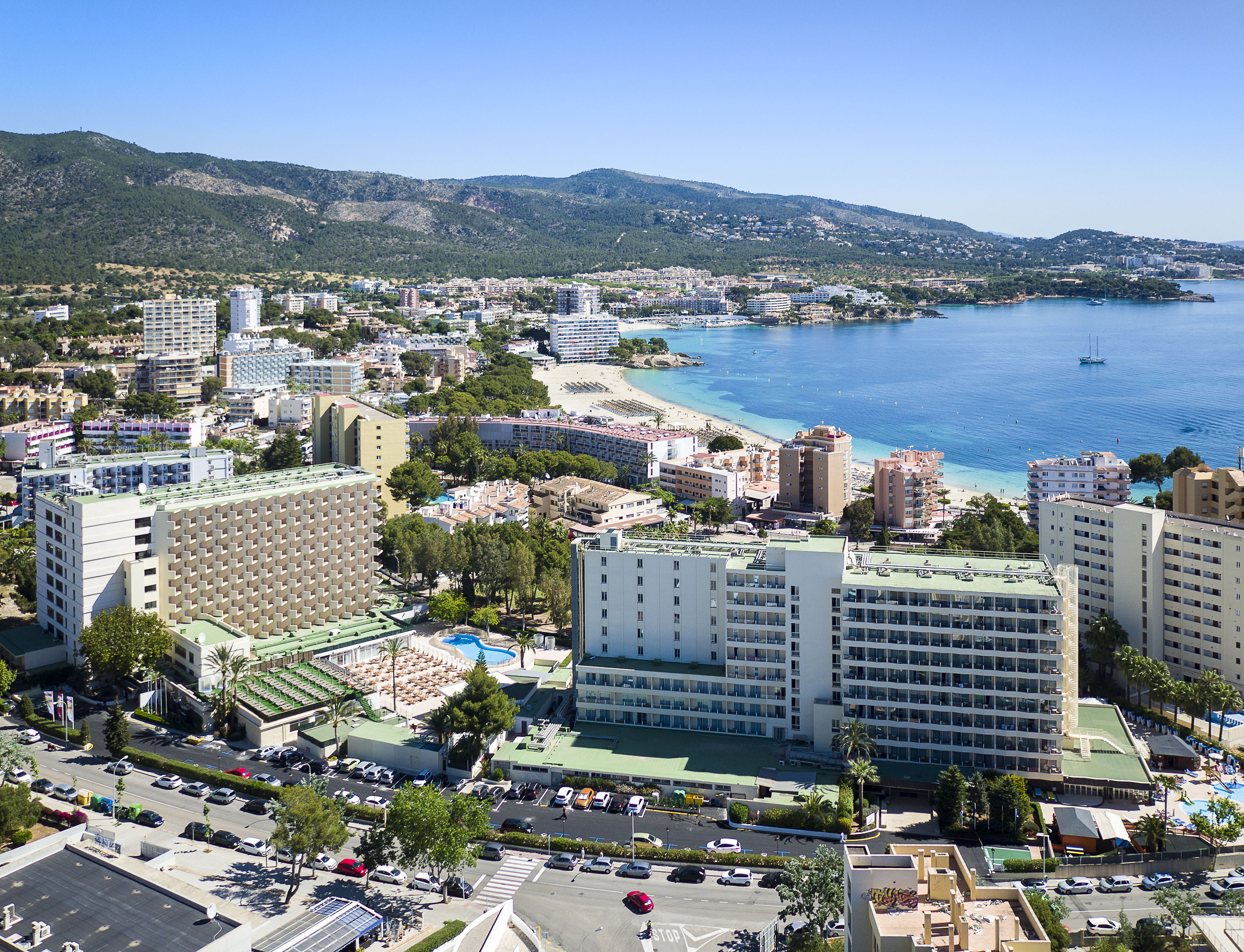 Sunny beach resort town with hotels, sandy beach, clear blue sea, and mountains.