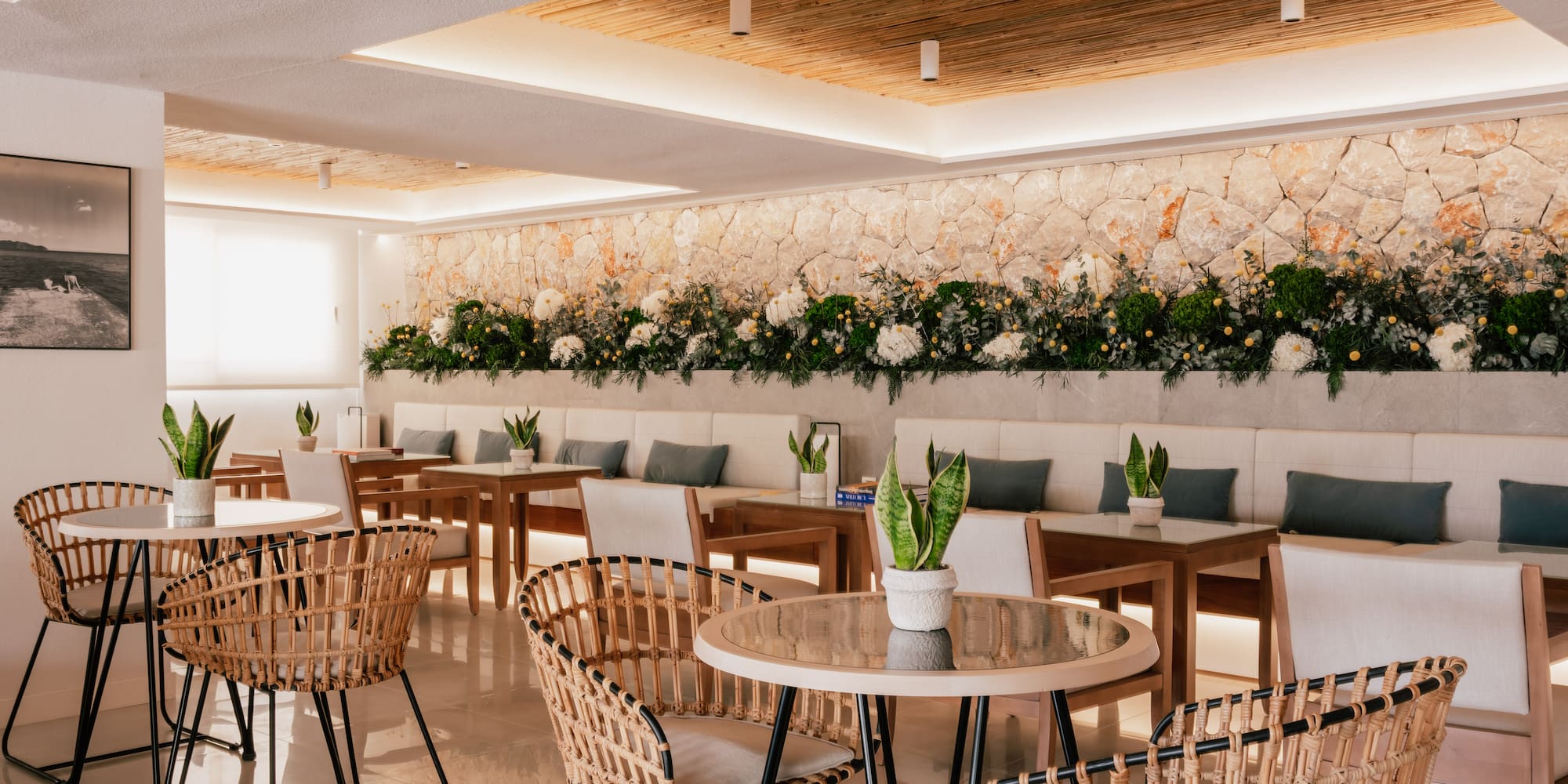 Modern cafe interior with wicker chairs, stone wall, and lush greenery.