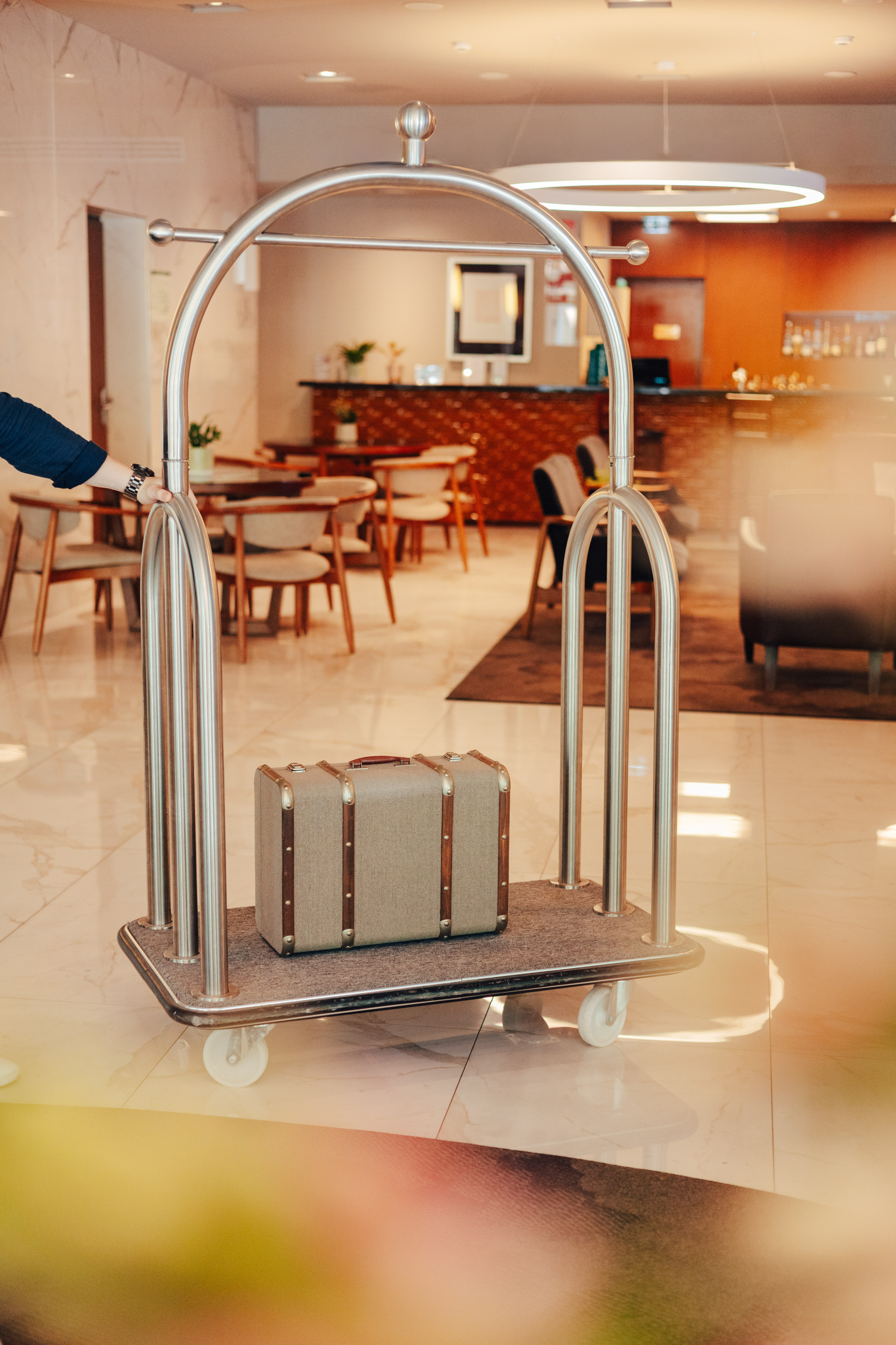 Hotel luggage cart with vintage suitcase pushed through modern lobby.