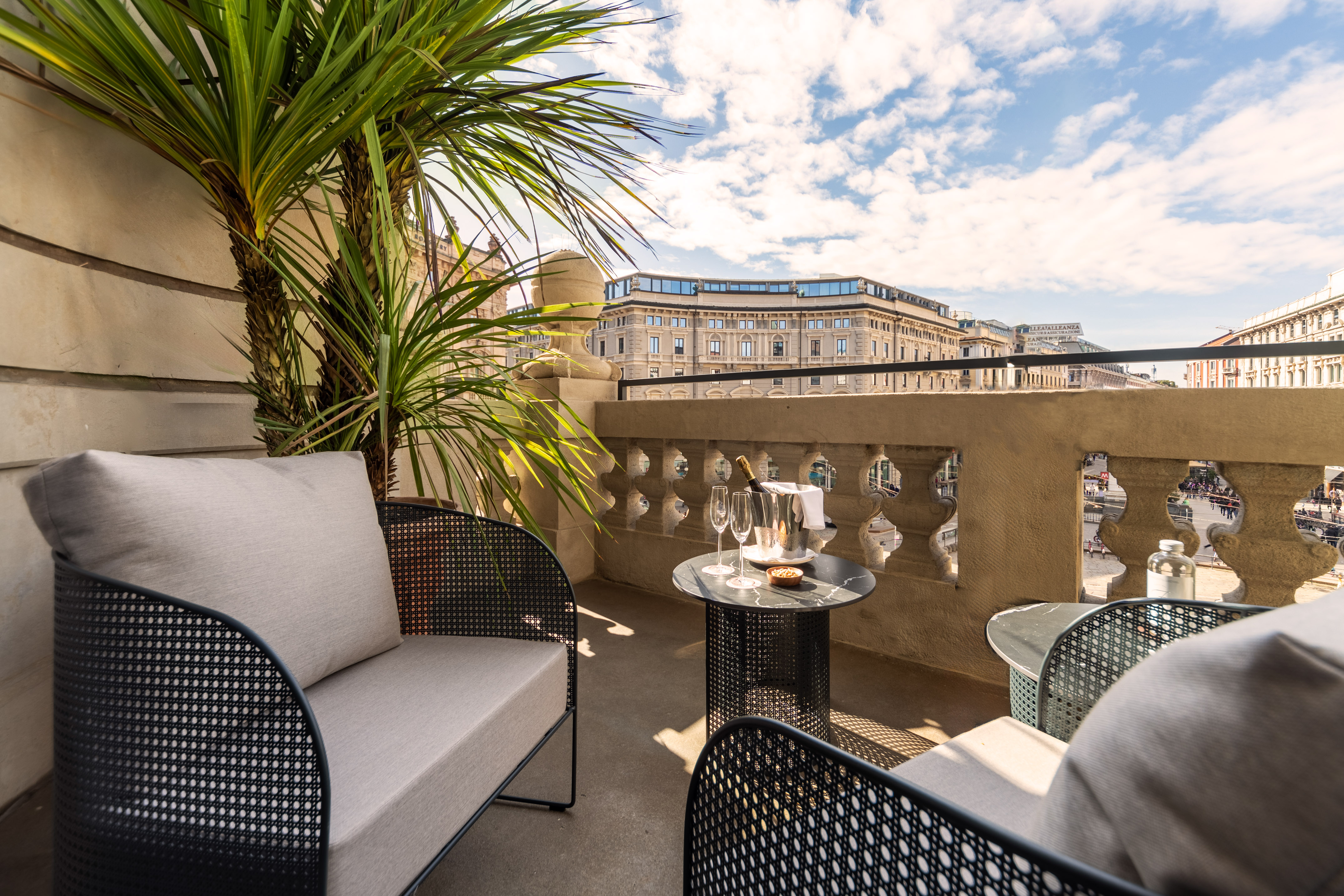 Hotel balcony with modern seating, champagne, palm plant, and city skyline.