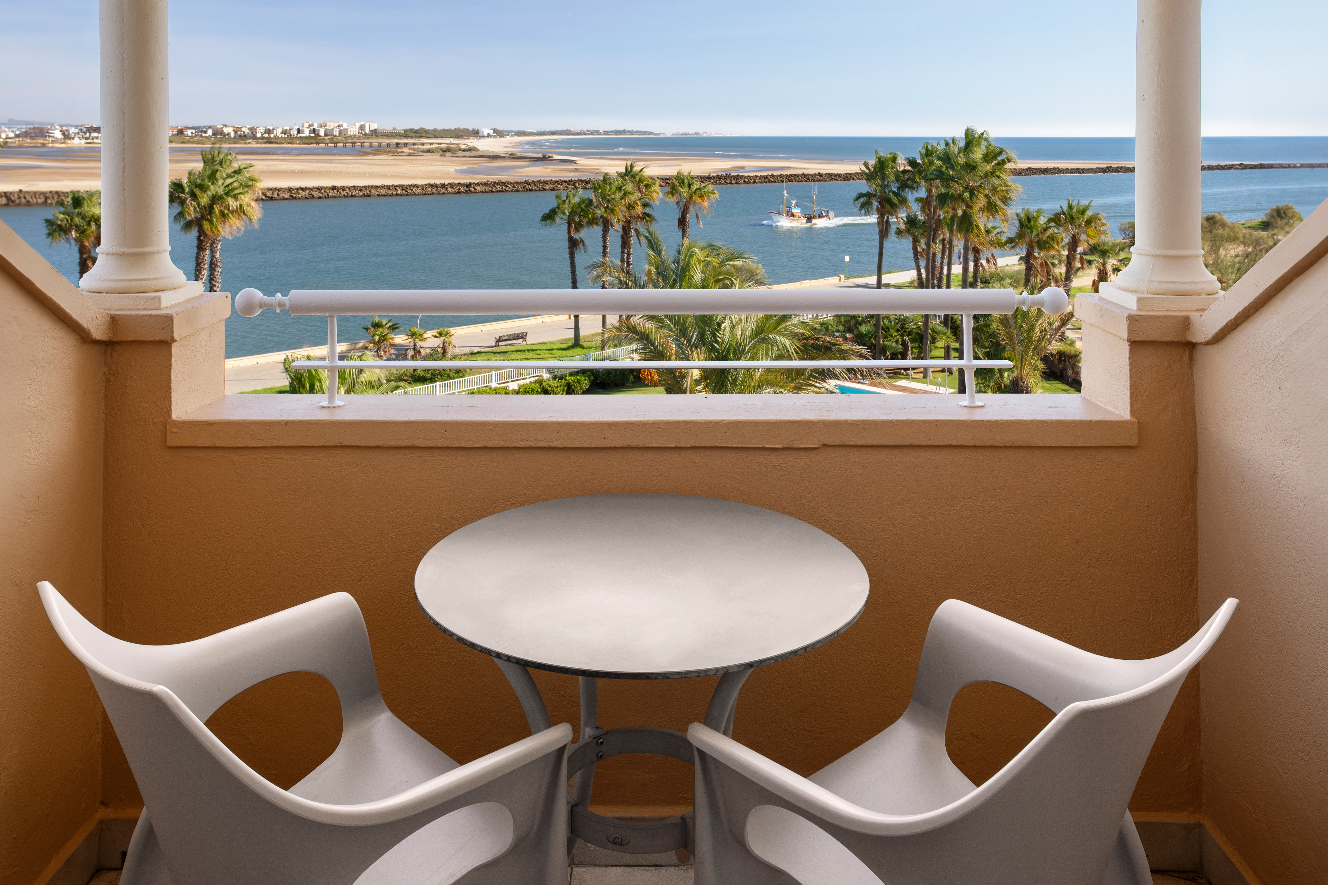 Resort balcony with seating, ocean view, palm trees, and boat.