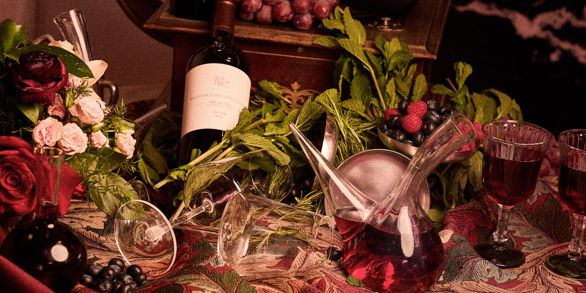 Dark red wine, decanter, glasses, grapes, berries, and roses on a rich cloth.