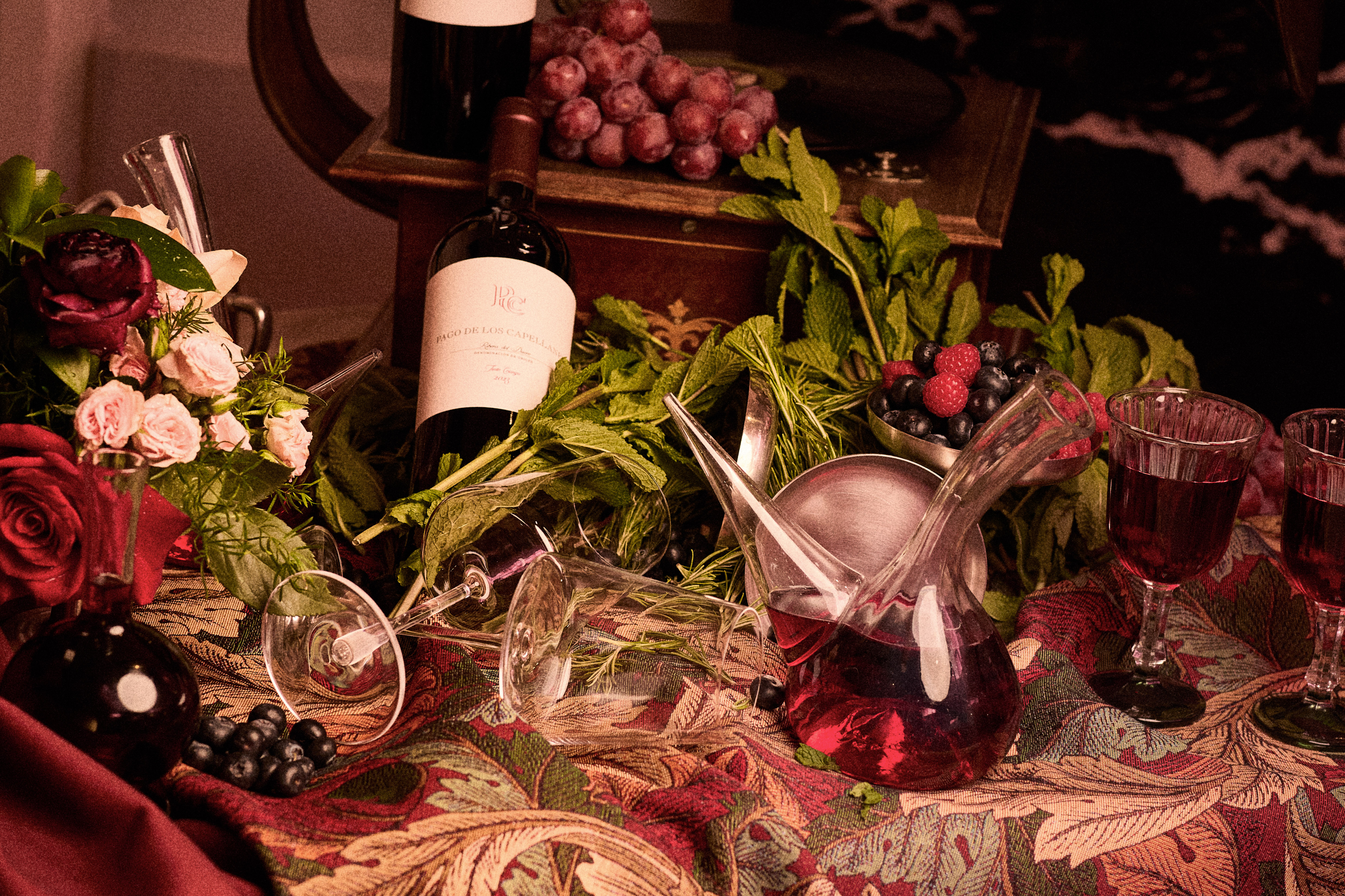 Dark red wine, decanter, glasses, grapes, berries, and roses on a rich cloth.