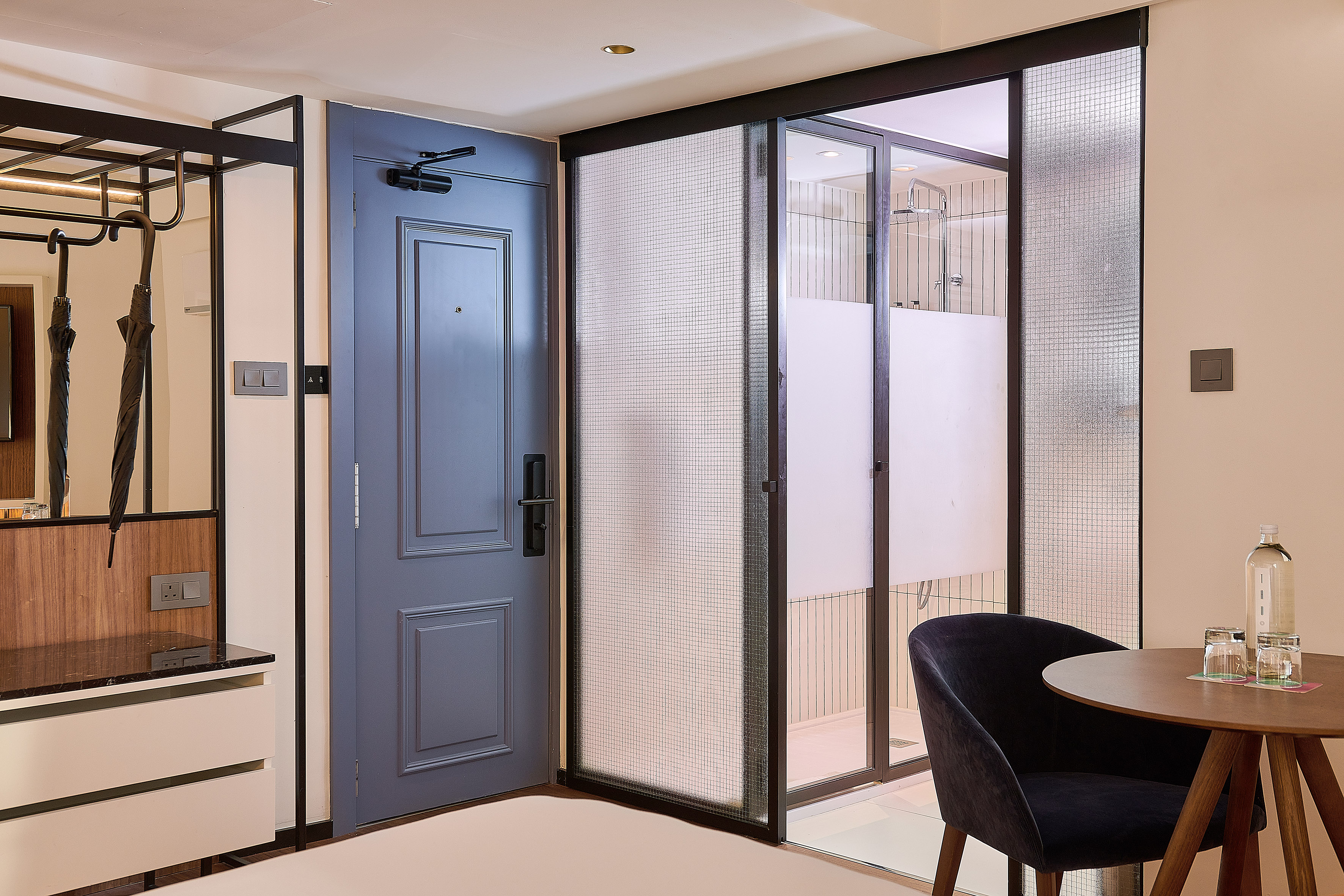 Hotel room interior with dark blue door, glass shower, wardrobe, and table.