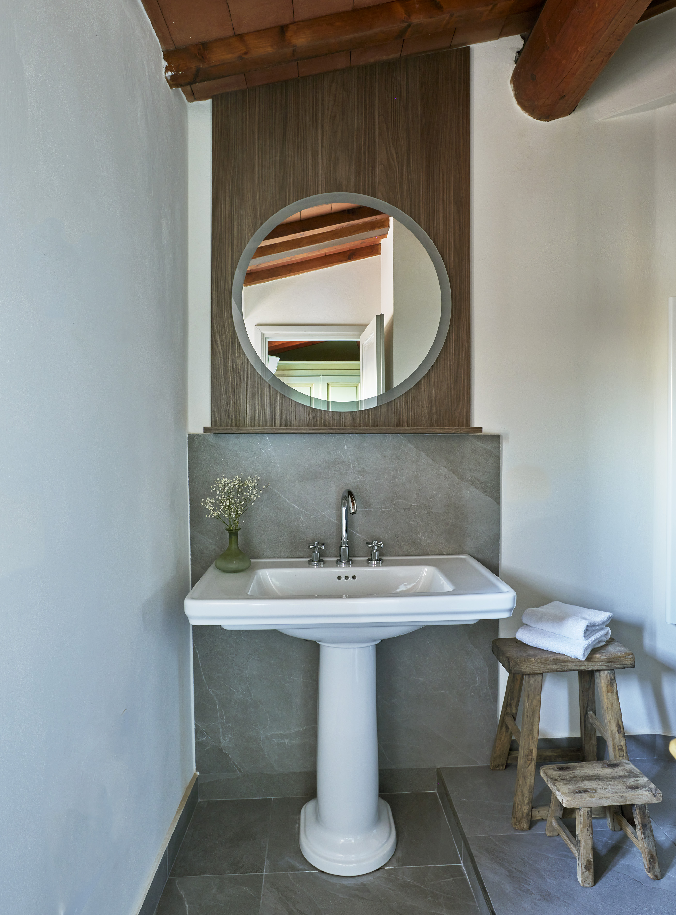 Modern bathroom with pedestal sink, round mirror, wooden beams, and rustic stool.