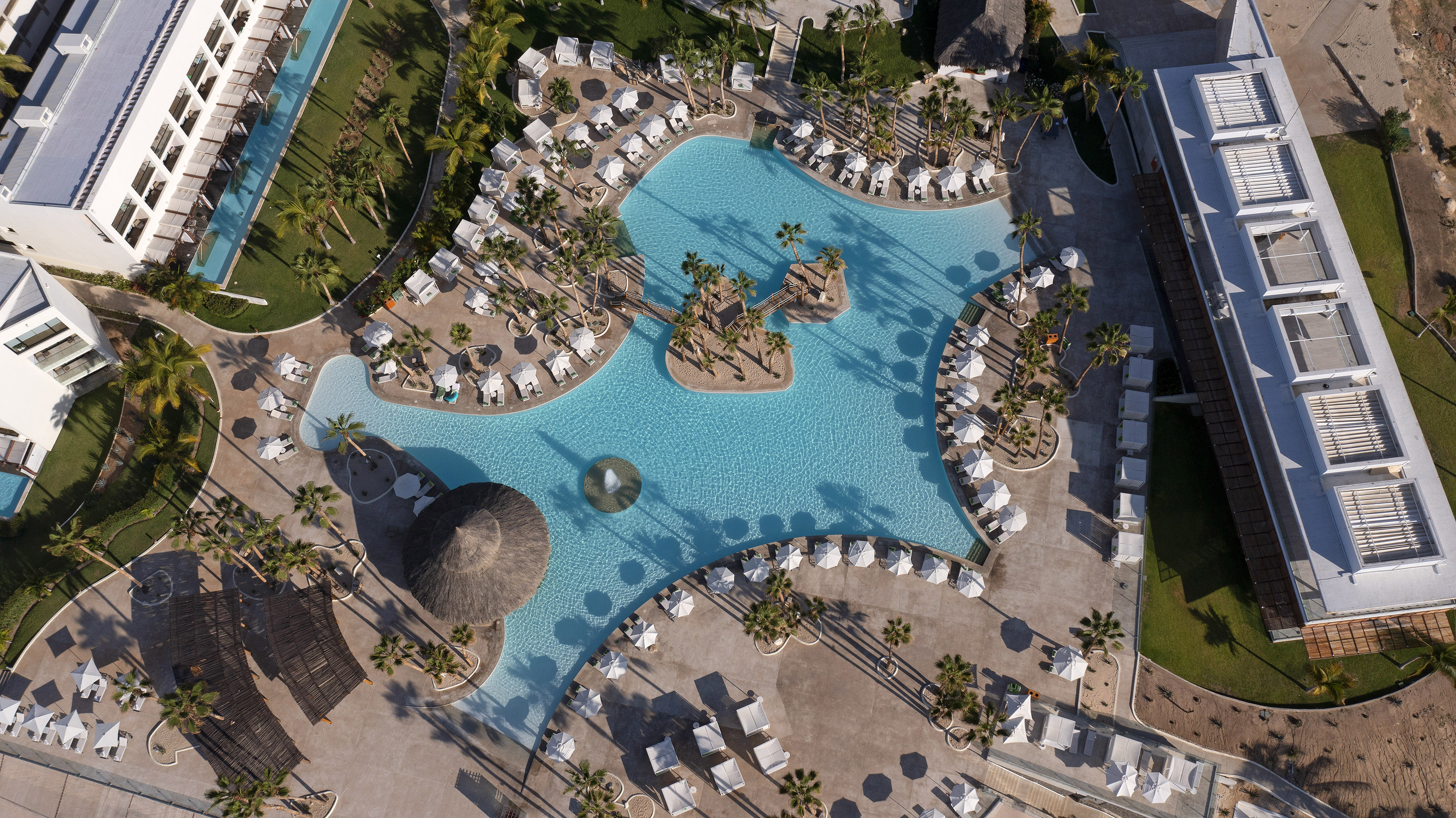Aerial: Luxury resort pool, island, palm trees, lounge chairs, modern buildings.