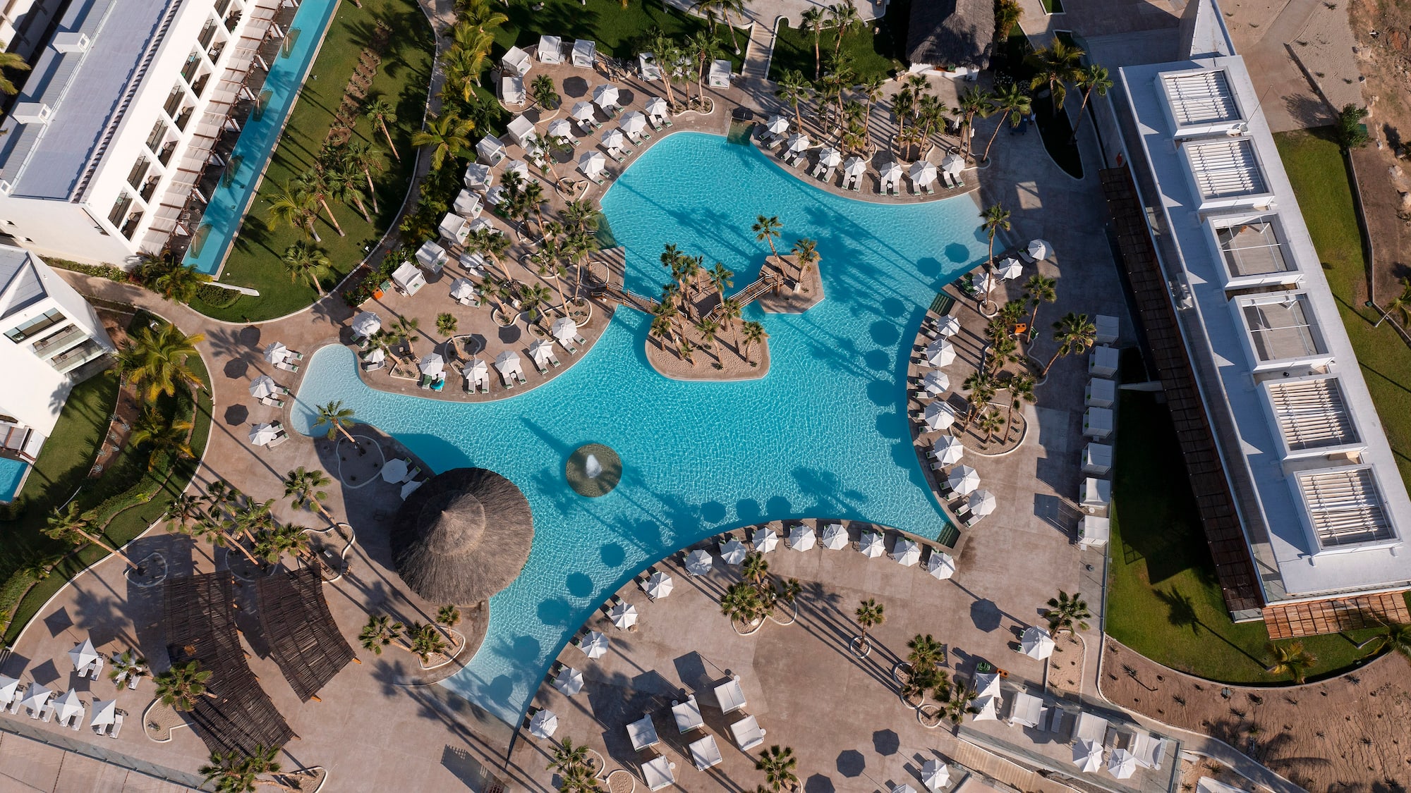 Aerial: Luxury resort pool, island, palm trees, lounge chairs, modern buildings.