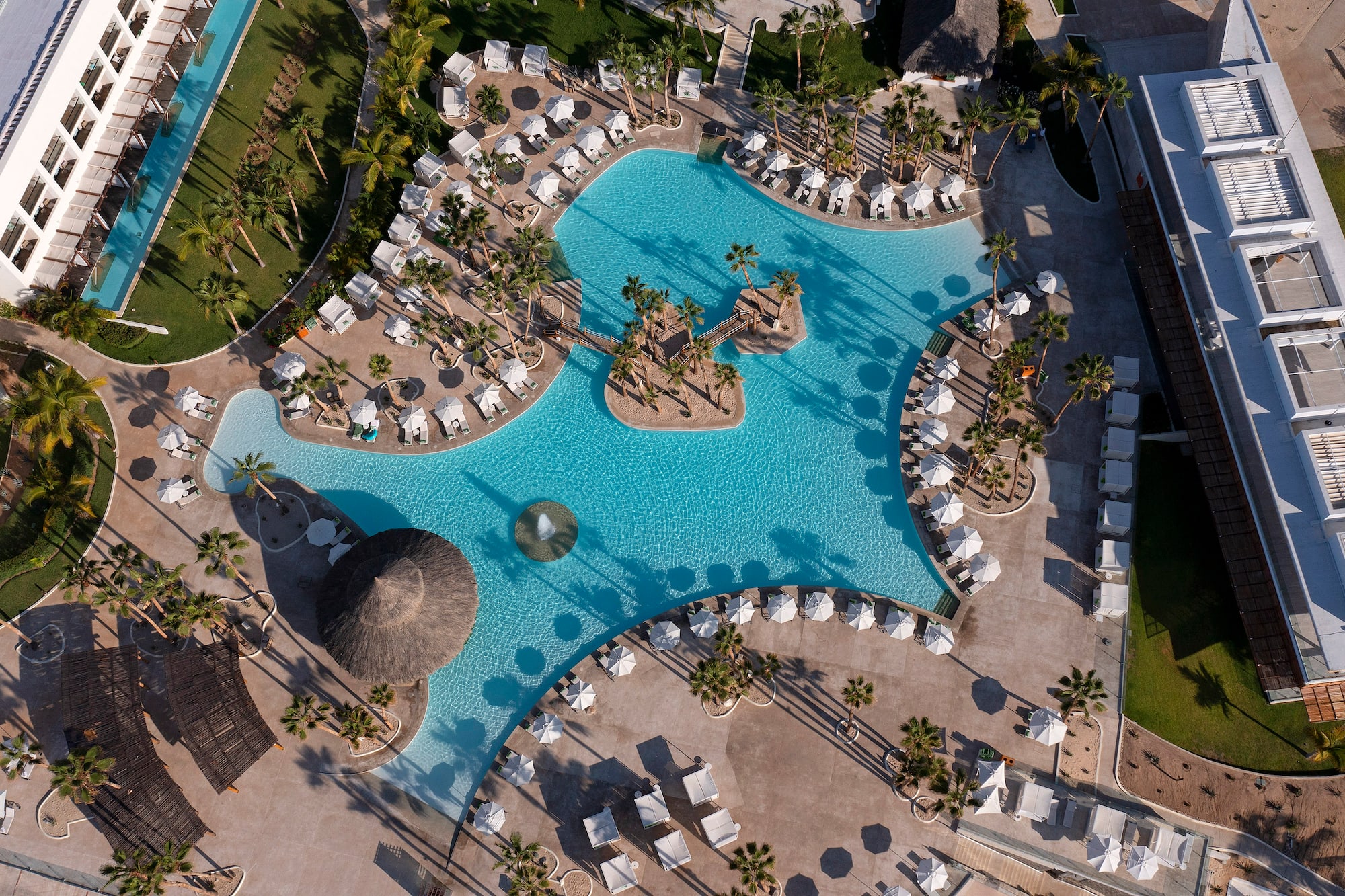 Aerial: Luxury resort pool, island, palm trees, lounge chairs, modern buildings.
