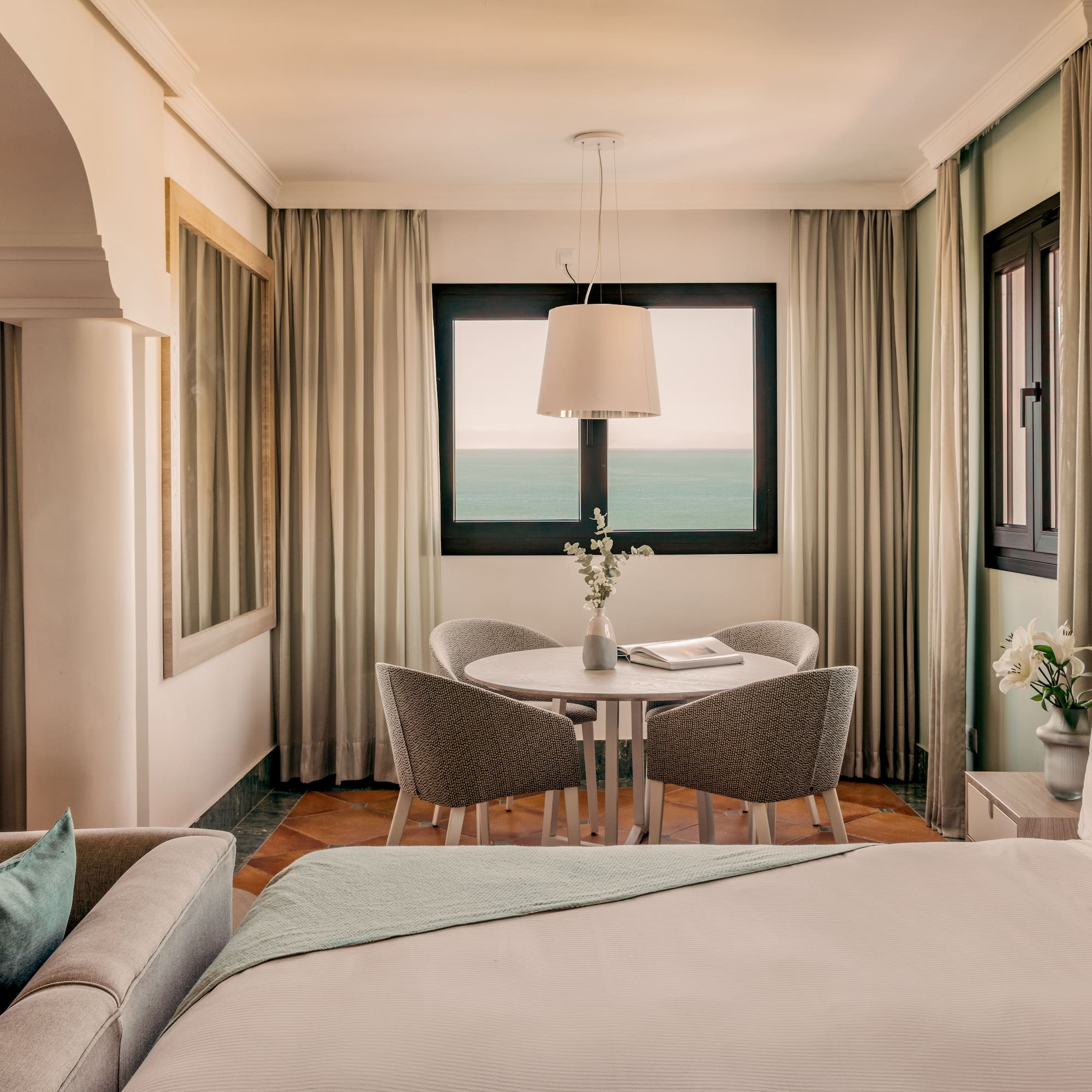 Hotel room with ocean view balcony, bed, sofa, and dining area.