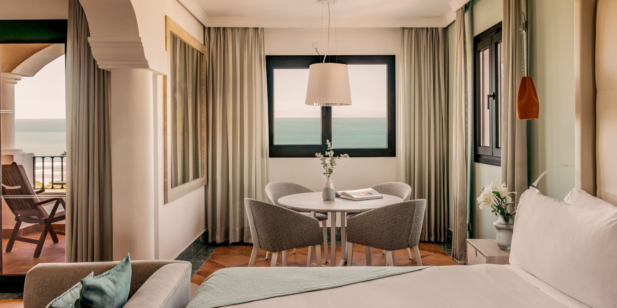 Hotel room with ocean view balcony, bed, sofa, and dining area.