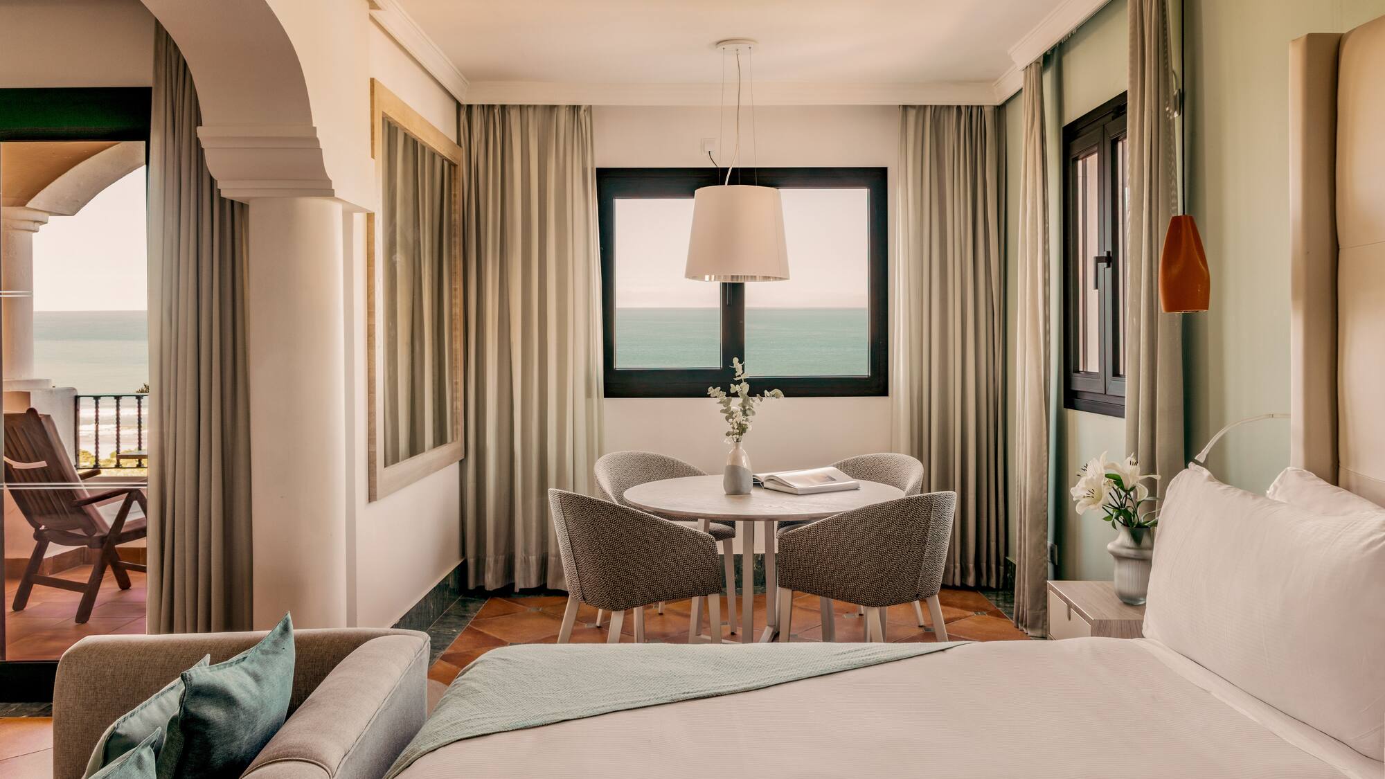 Hotel room with ocean view balcony, bed, sofa, and dining area.