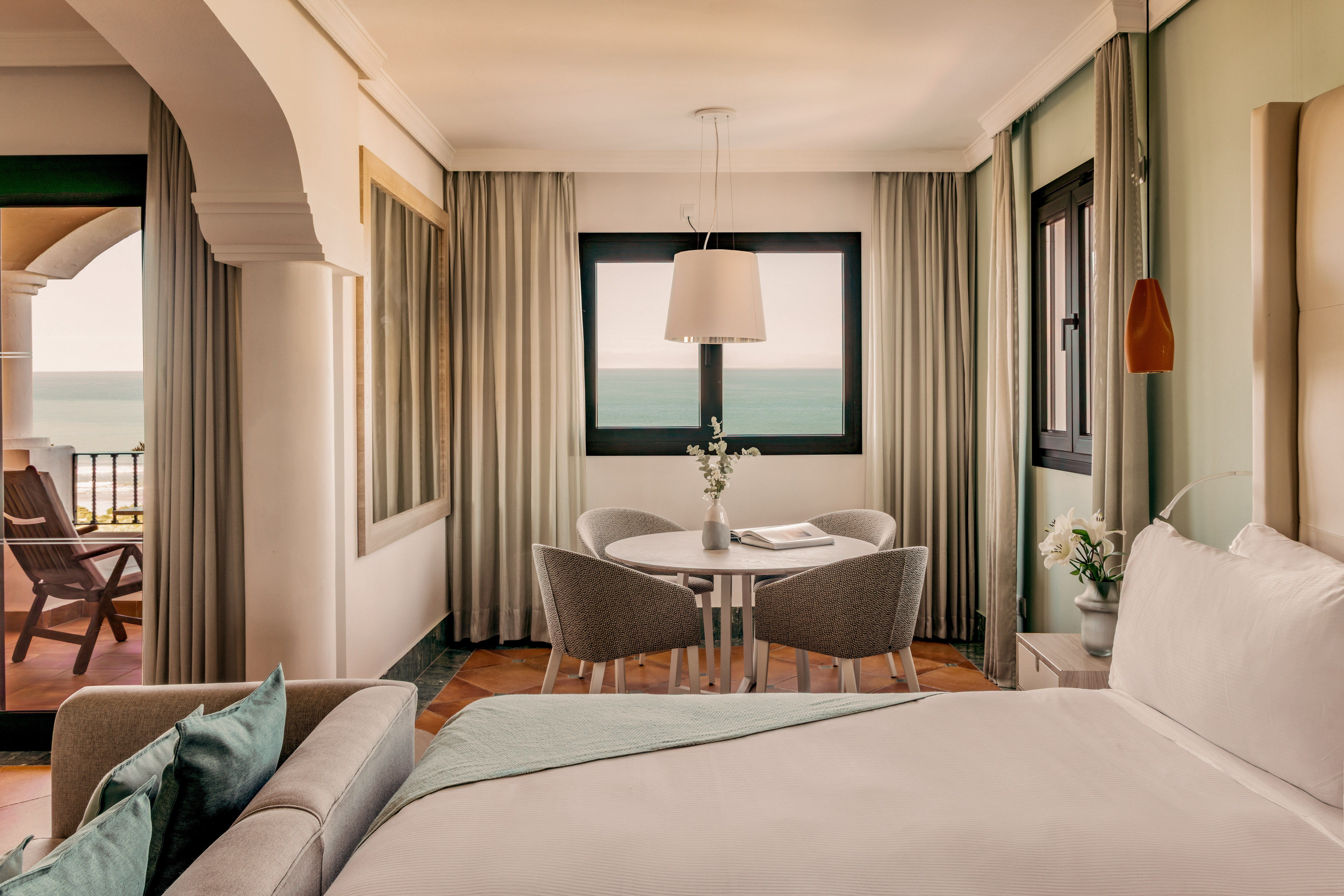 Hotel room with ocean view balcony, bed, sofa, and dining area.