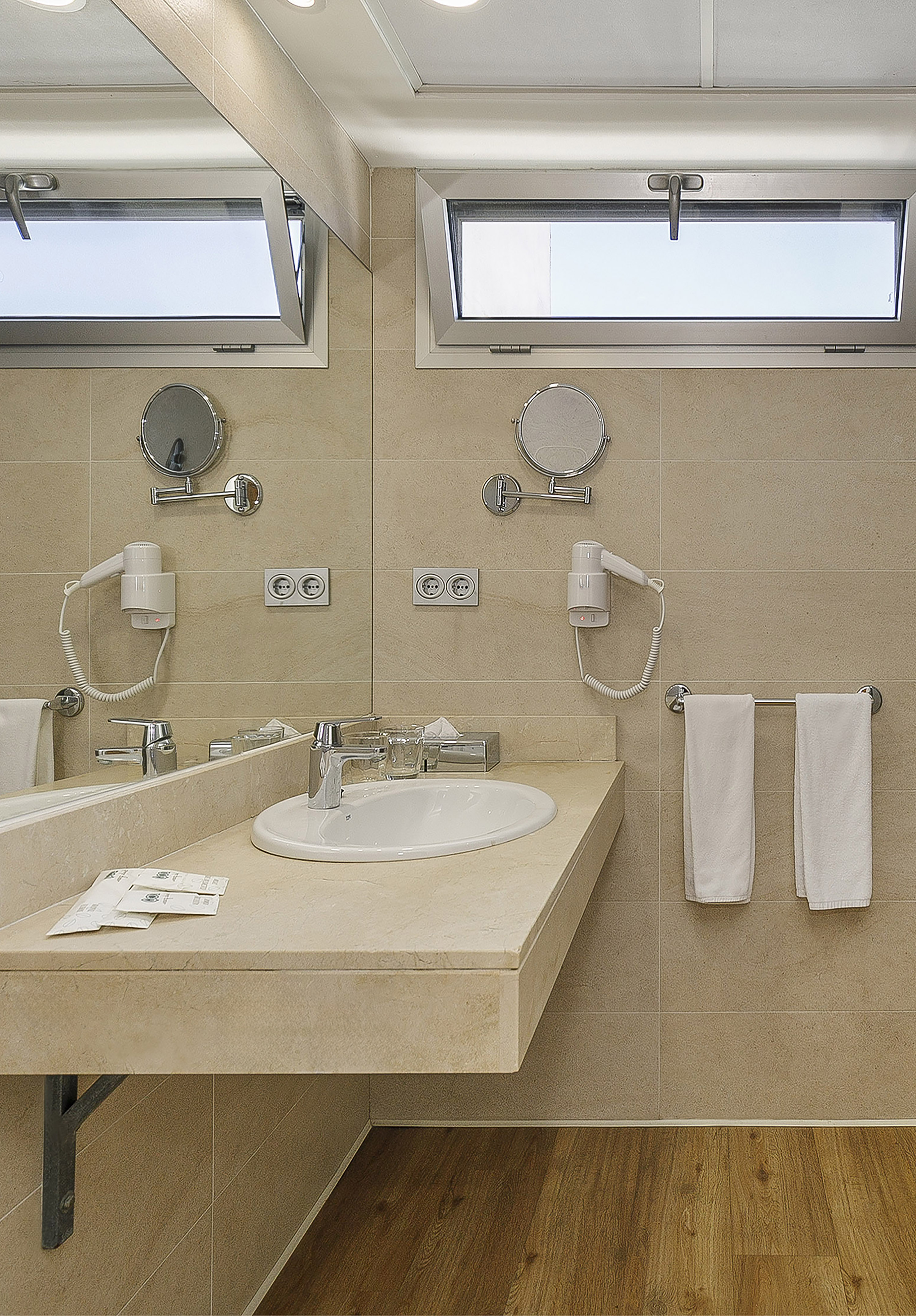 Modern hotel bathroom with beige vanity, white sink, windows, and hanging towels.
