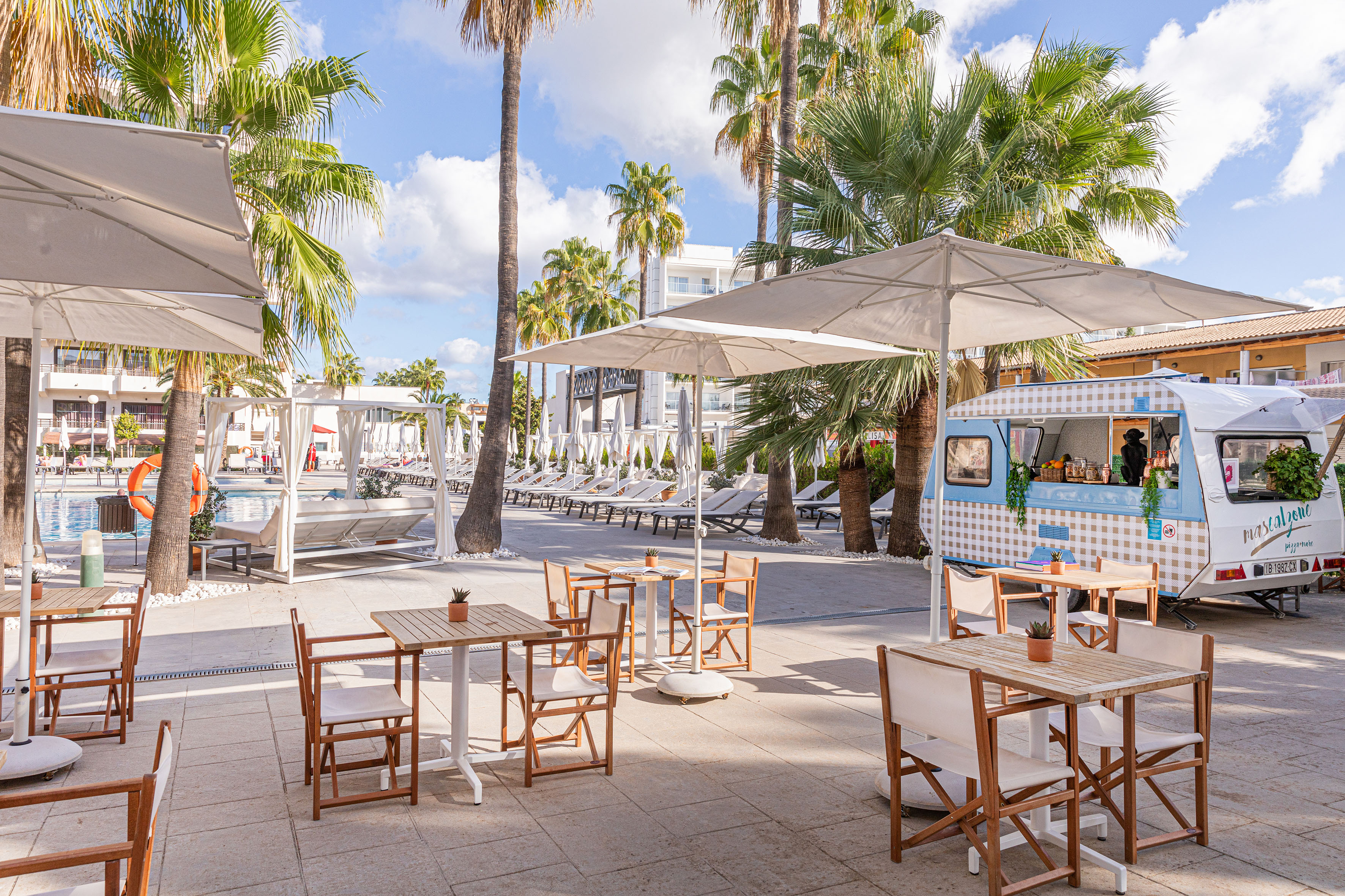 Sunny resort patio with outdoor dining, food truck, pool, and palm trees.