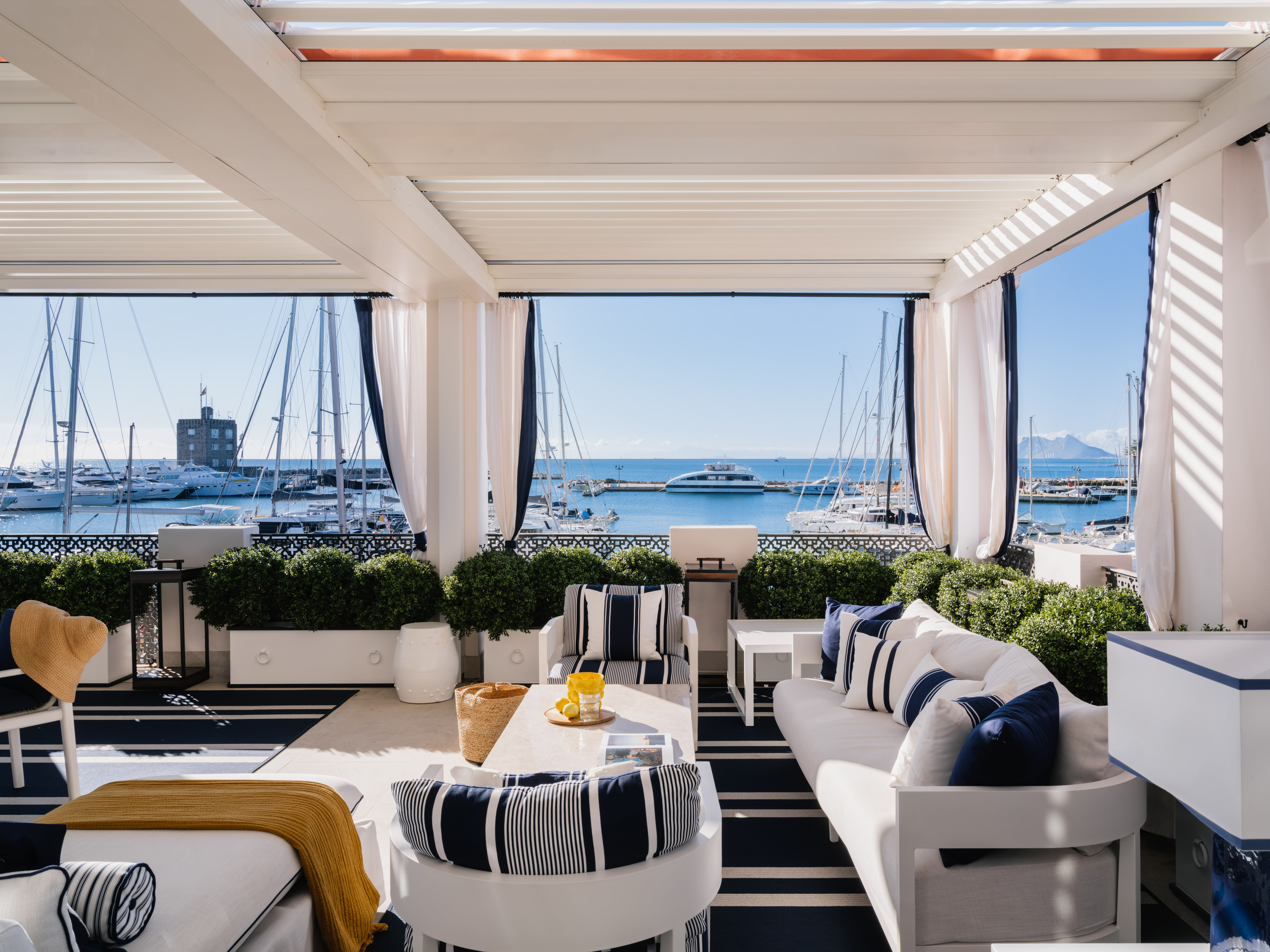 Waterfront patio with white pergola, striped nautical decor, overlooking a busy marina.