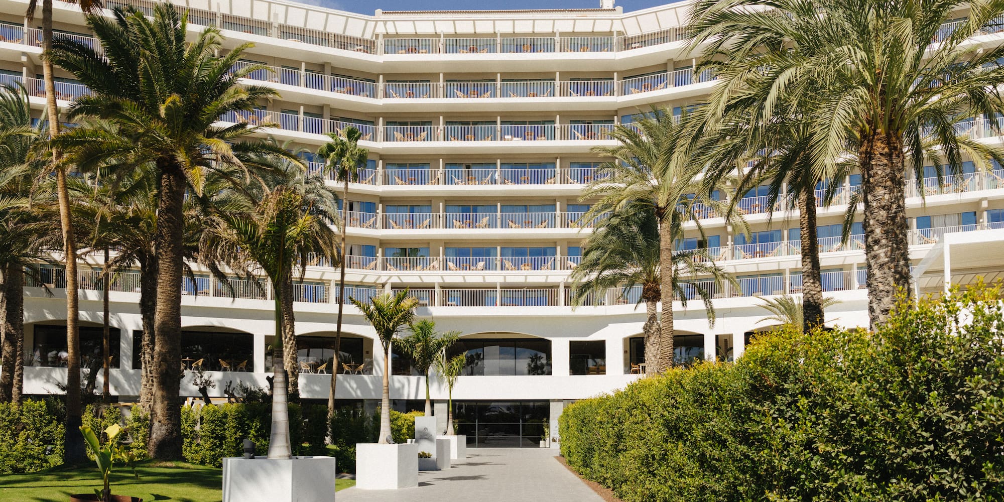 White curved resort hotel with numerous balconies, surrounded by palm trees.