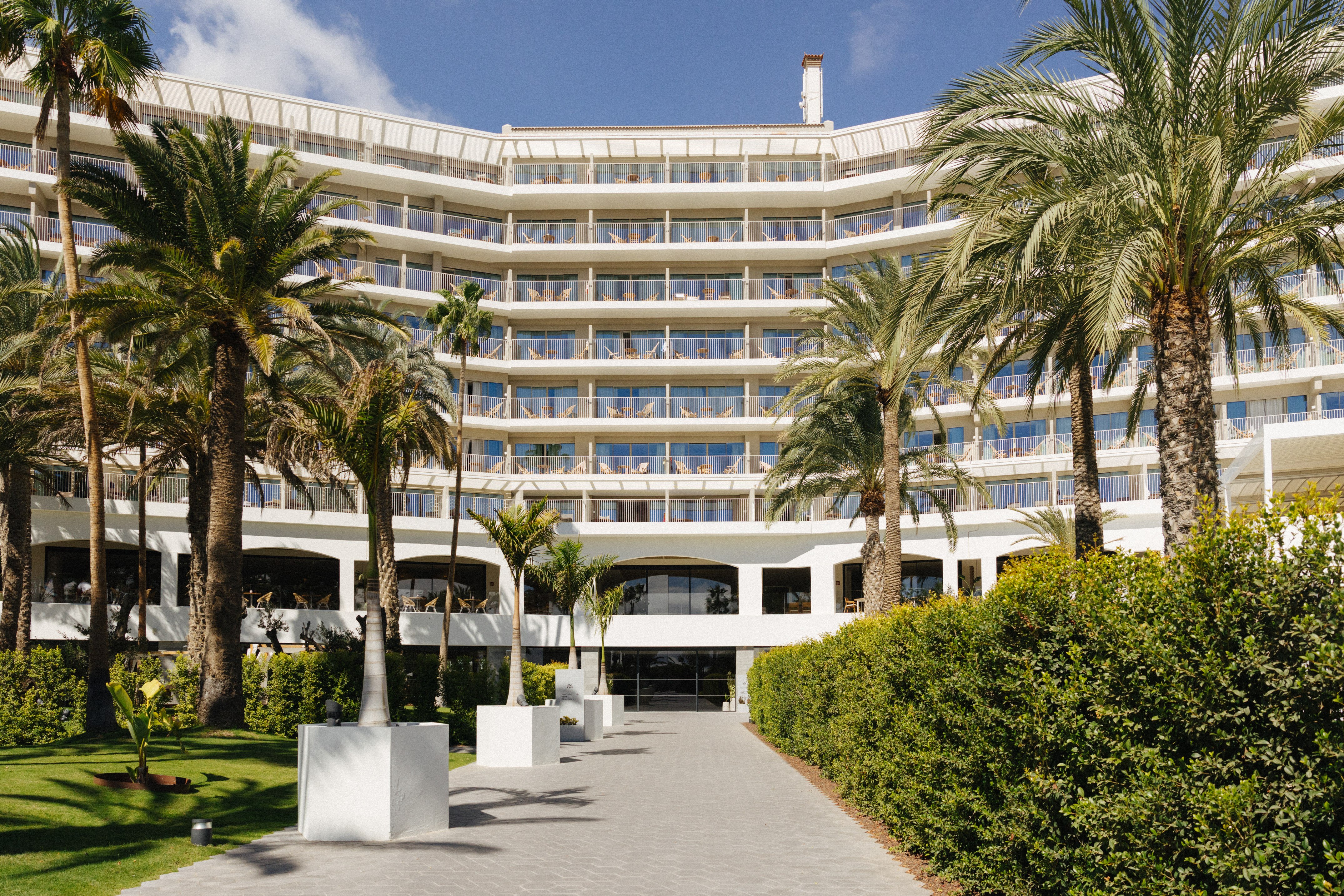 White curved resort hotel with numerous balconies, surrounded by palm trees.