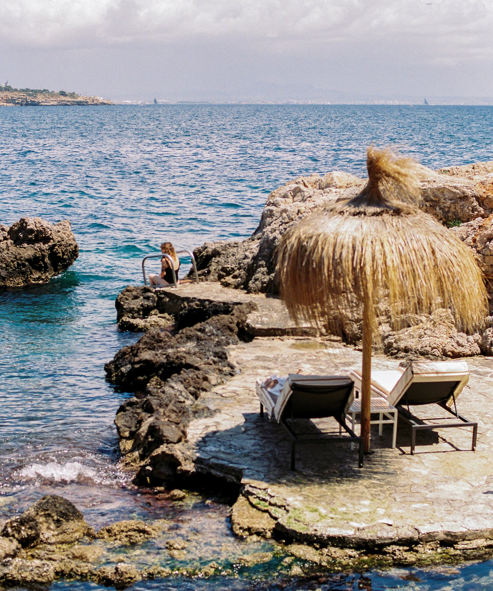 Rocky coastal retreat with person by clear sea, straw umbrella, sunbeds.