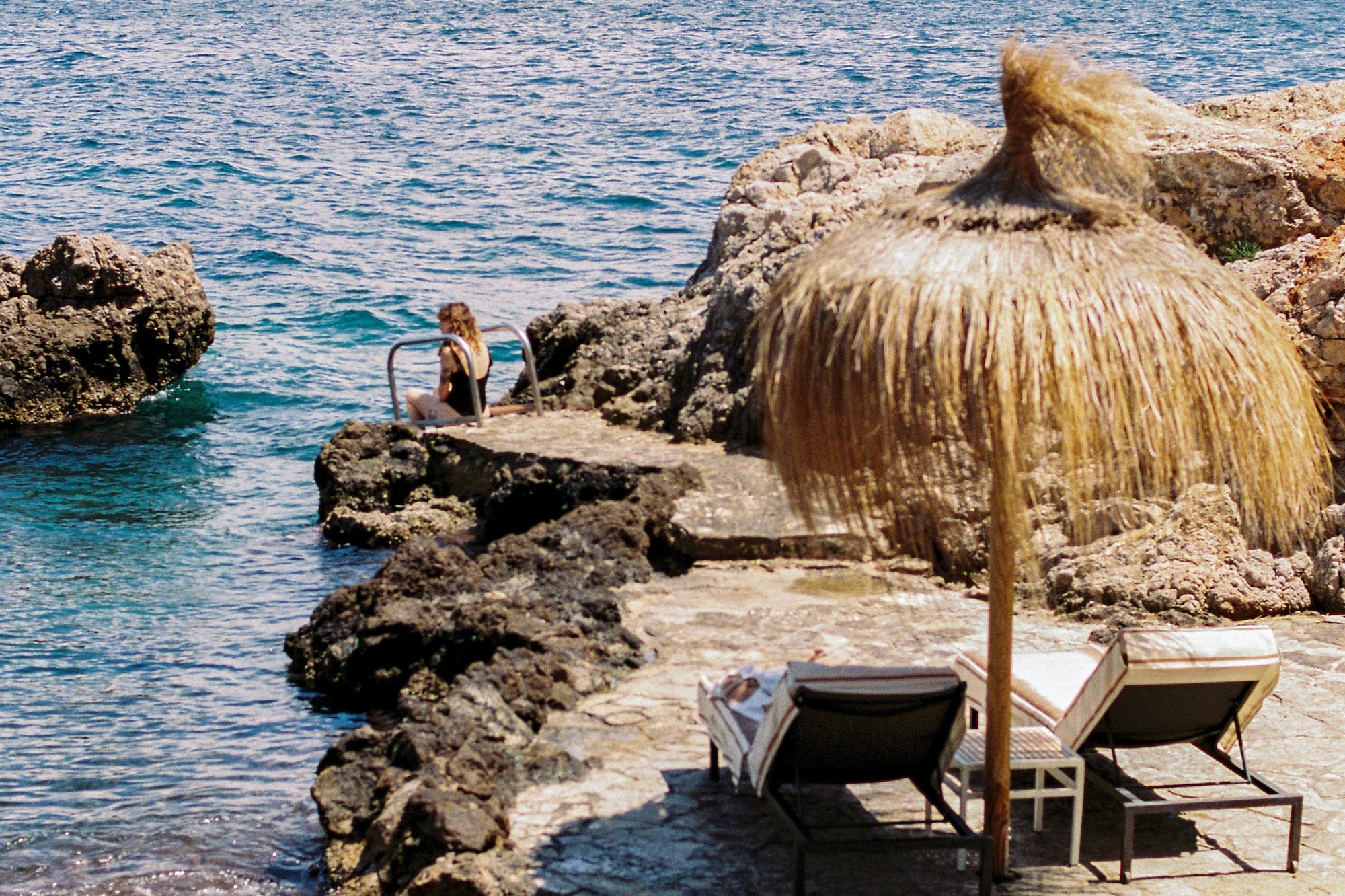 Rocky coastal retreat with person by clear sea, straw umbrella, sunbeds.