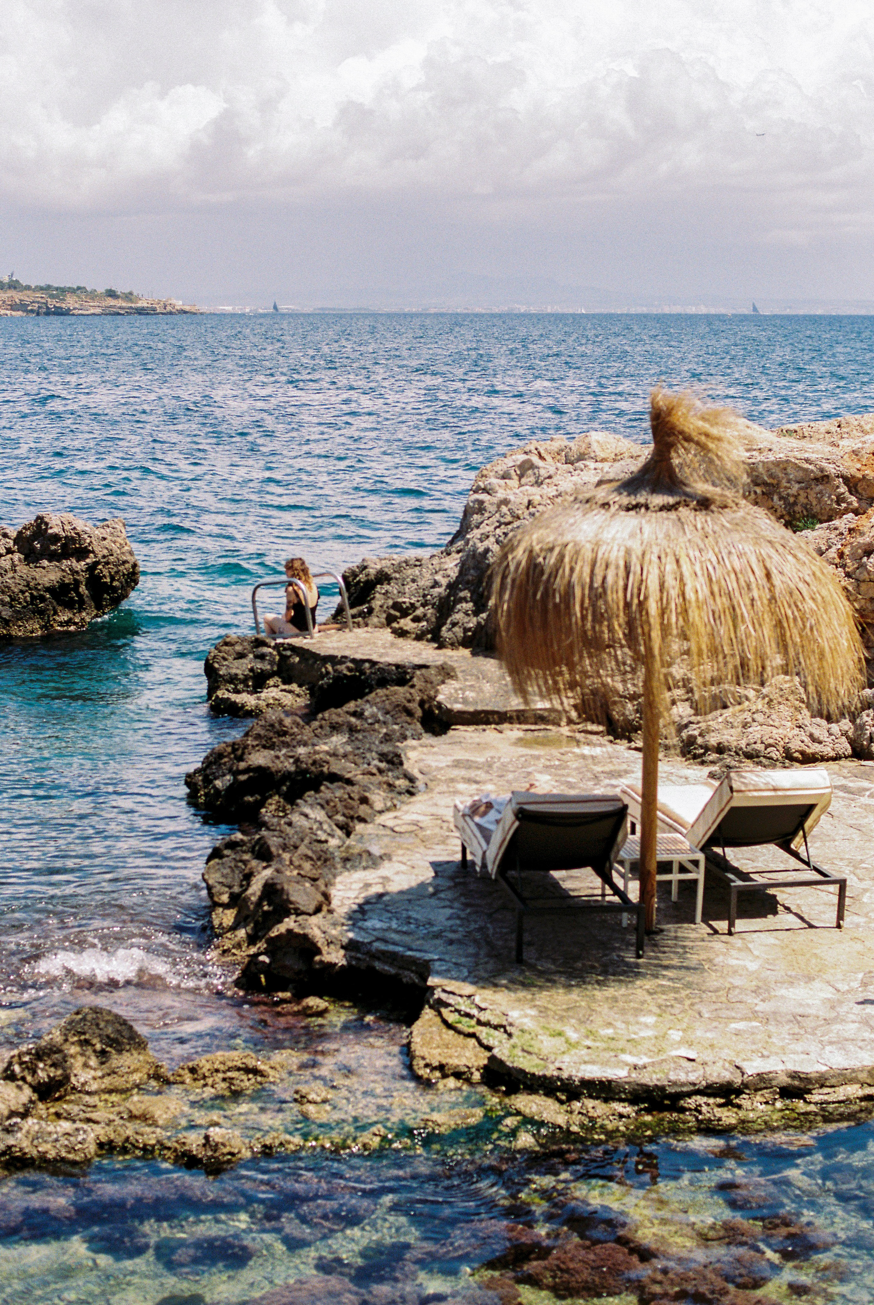 Rocky coastal retreat with person by clear sea, straw umbrella, sunbeds.