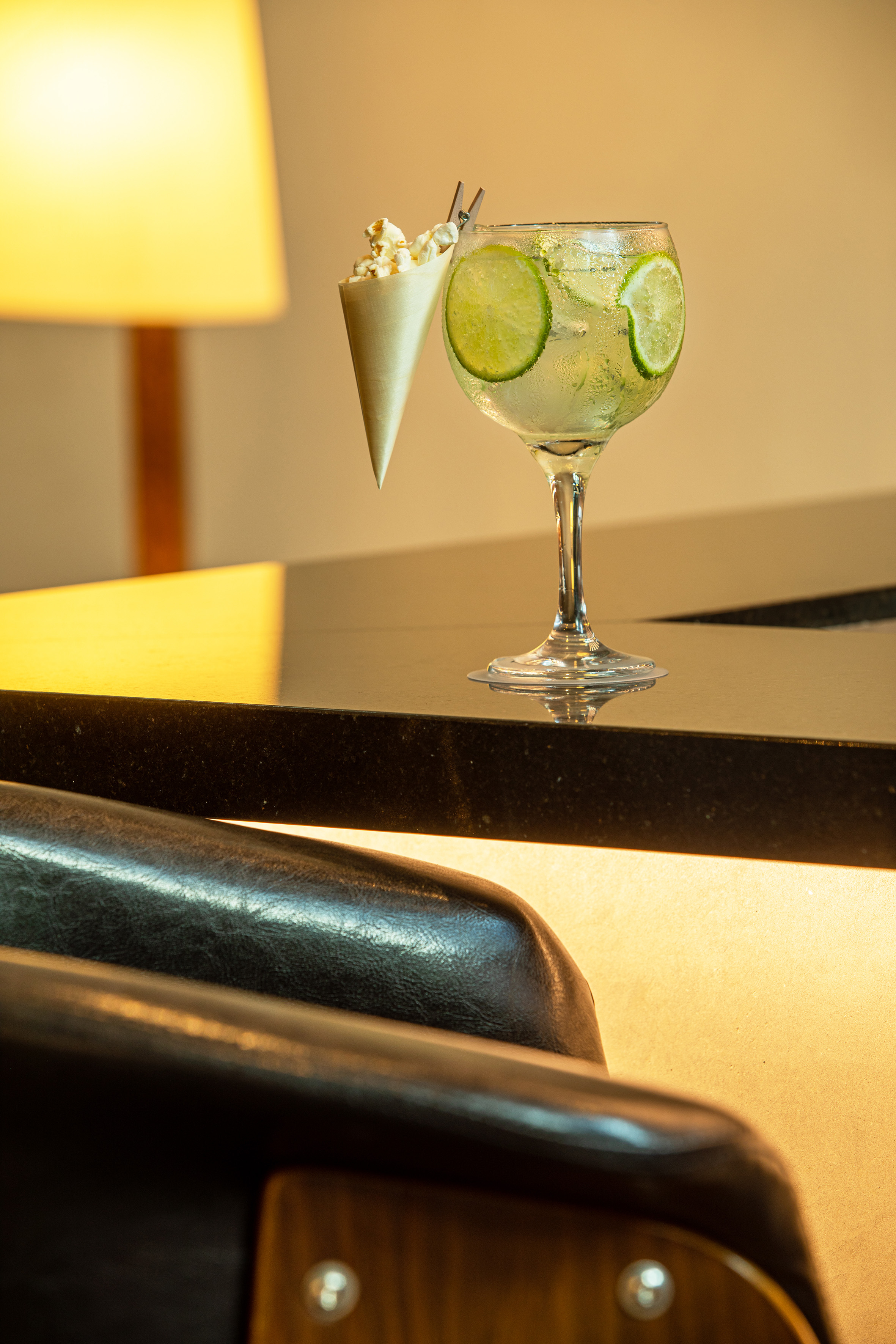 Gin and tonic cocktail with lime slices and popcorn cone on a dark bar table.
