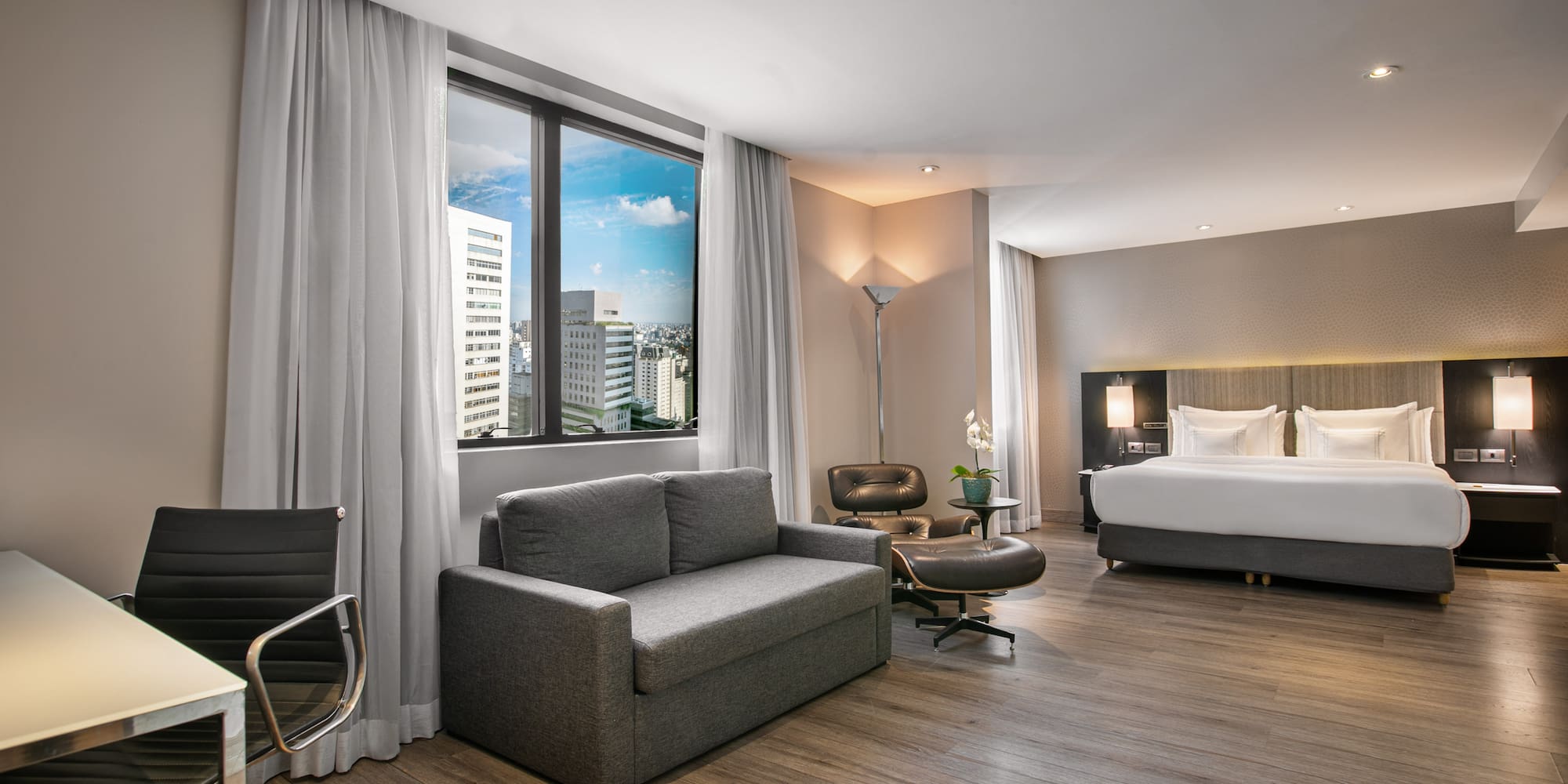 Modern hotel suite with city view, king bed, sofa bed, and desk.