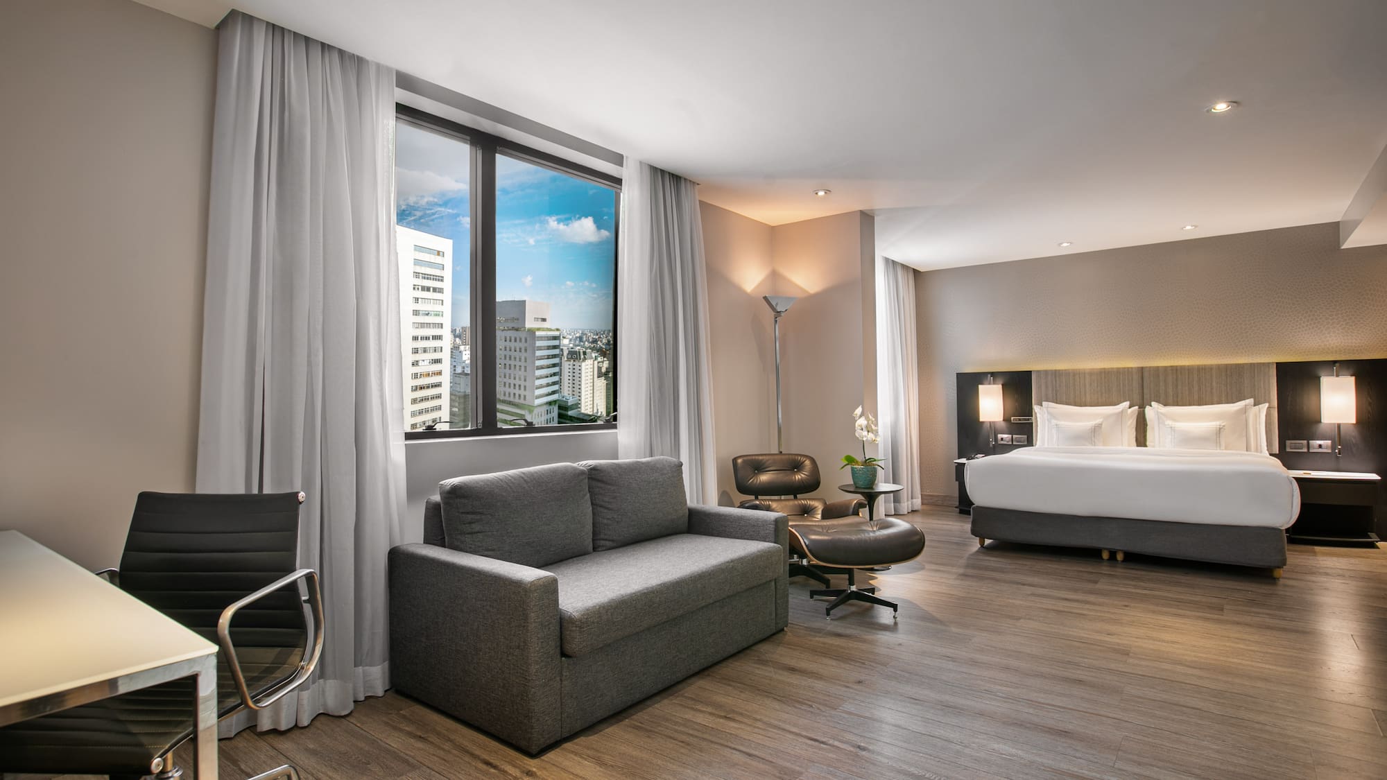 Modern hotel suite with city view, king bed, sofa bed, and desk.