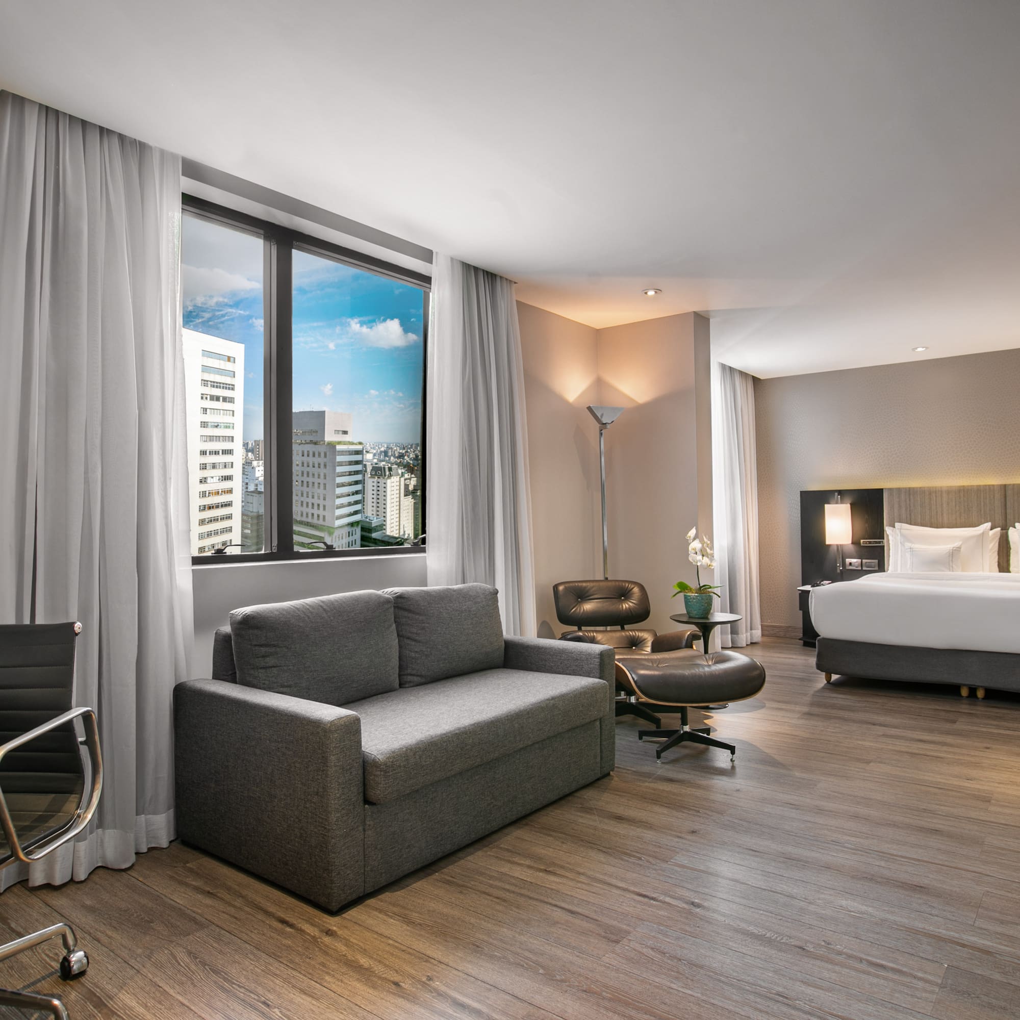 Modern hotel suite with city view, king bed, sofa bed, and desk.