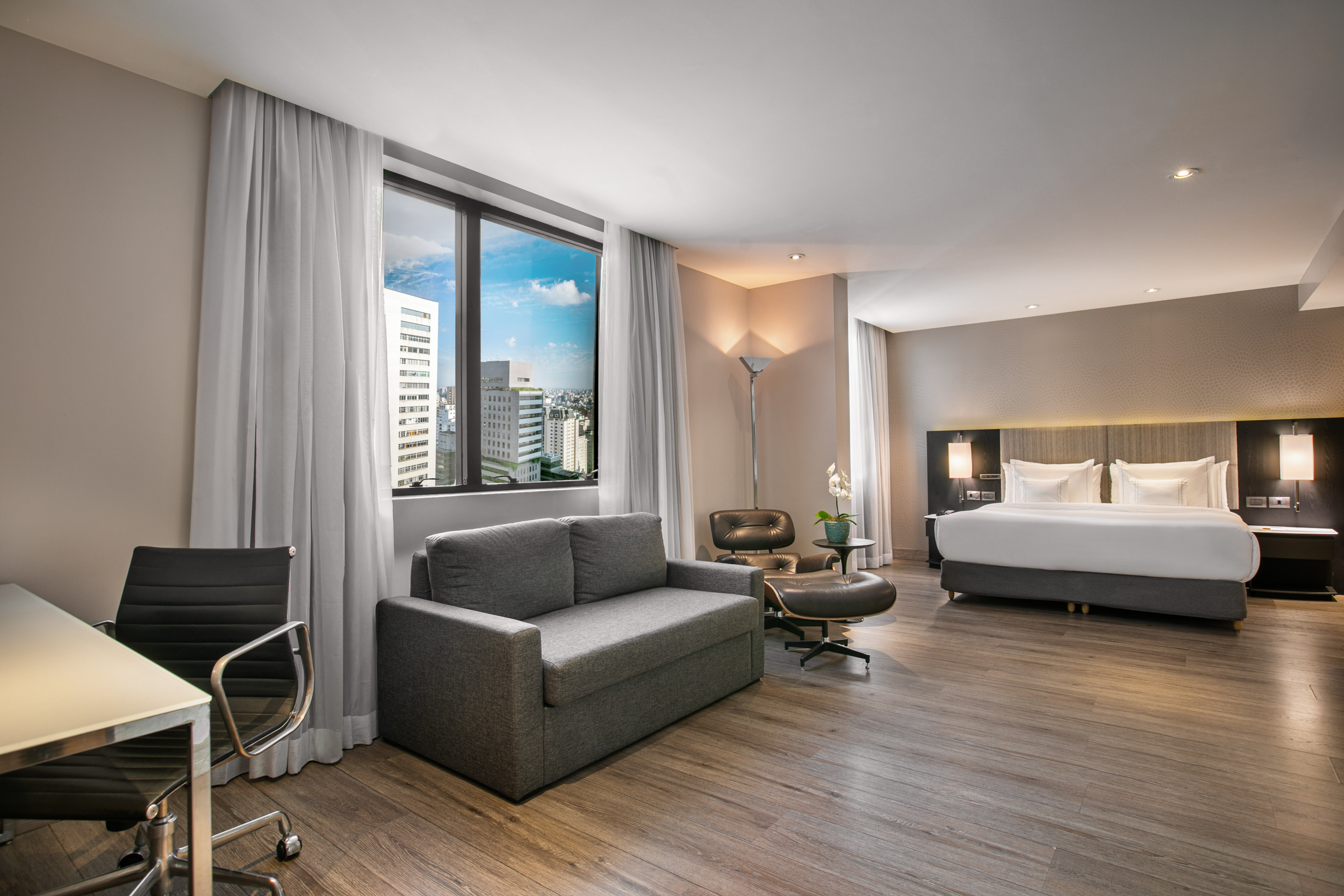 Modern hotel suite with city view, king bed, sofa bed, and desk.
