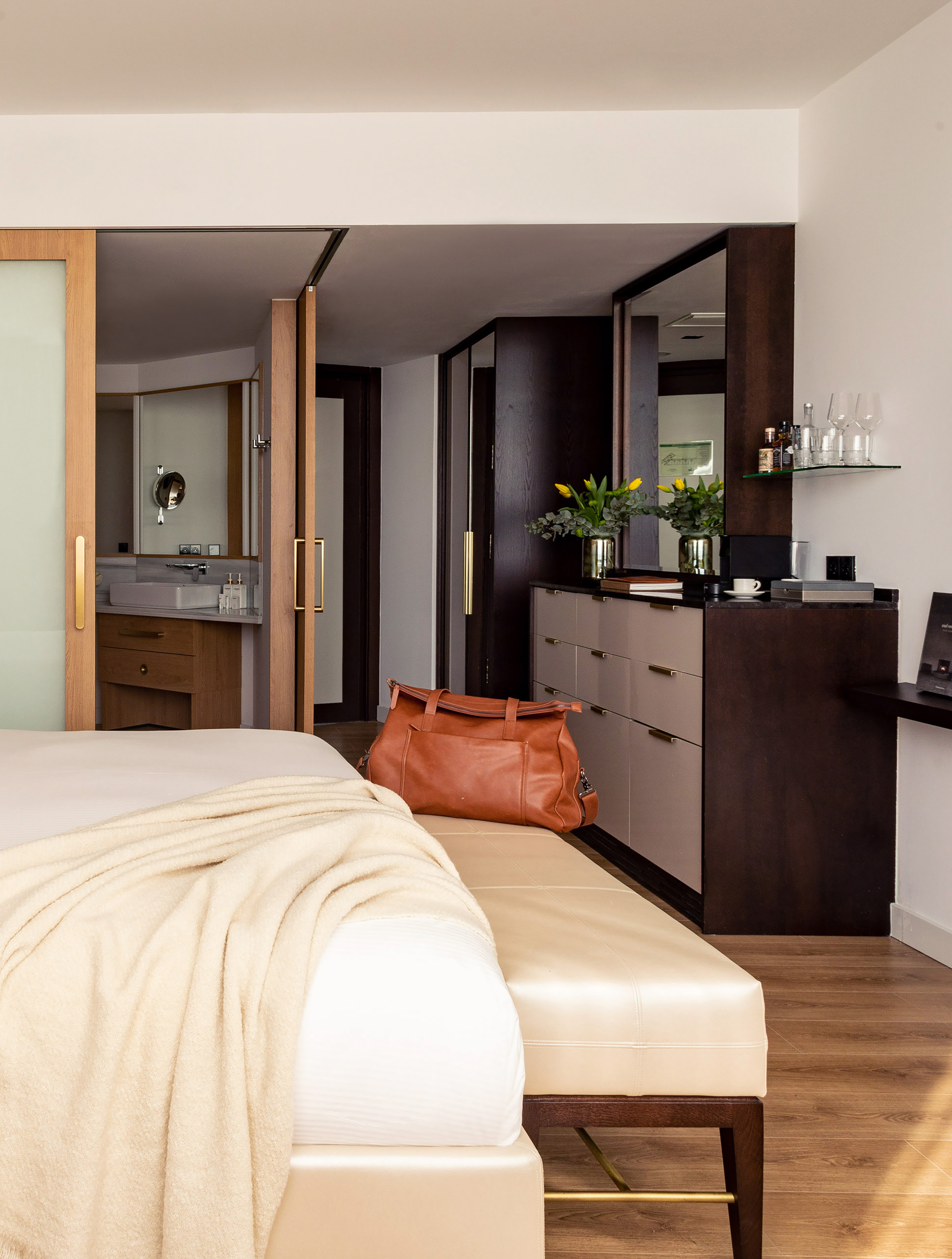 Modern hotel bedroom with bed, beige bench, leather travel bag, and dresser.