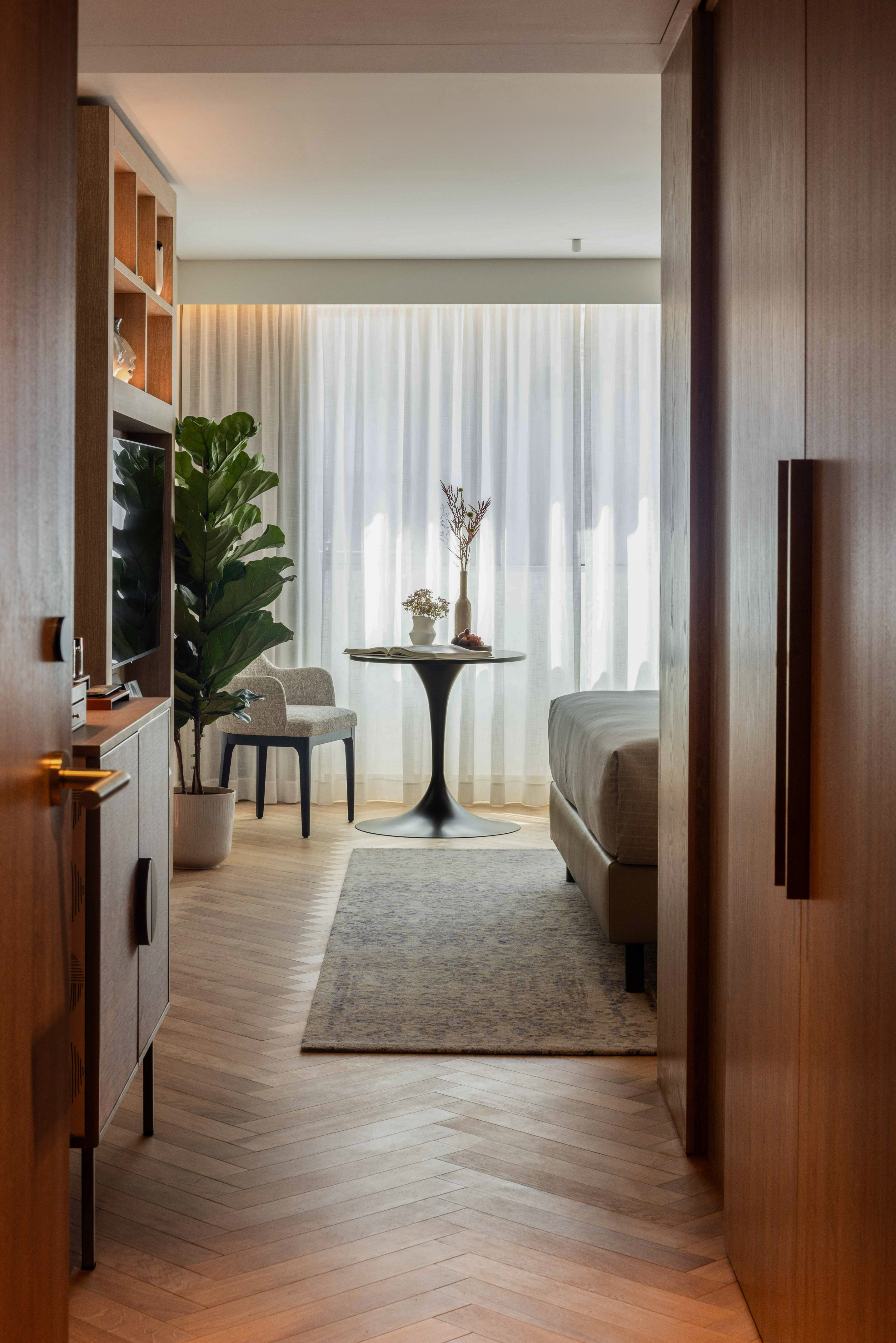 Modern hotel room entryway with wood flooring, bed, plant, and sheer curtains.