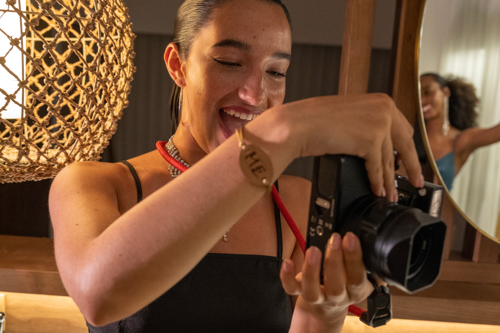 Laughing woman in black dress holding camera, another woman in mirror.