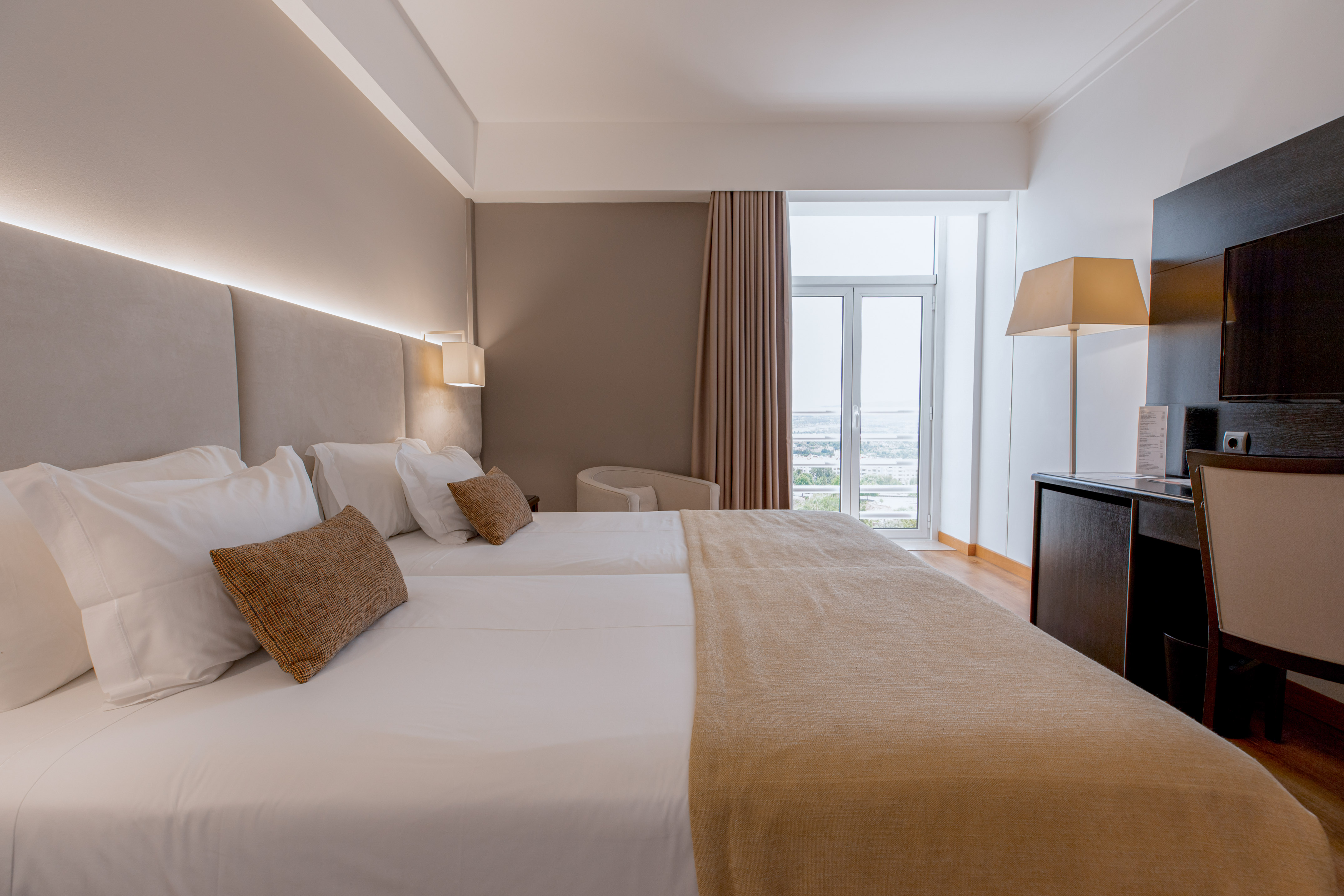 Modern hotel room with twin beds, padded headboard, desk, and window view.