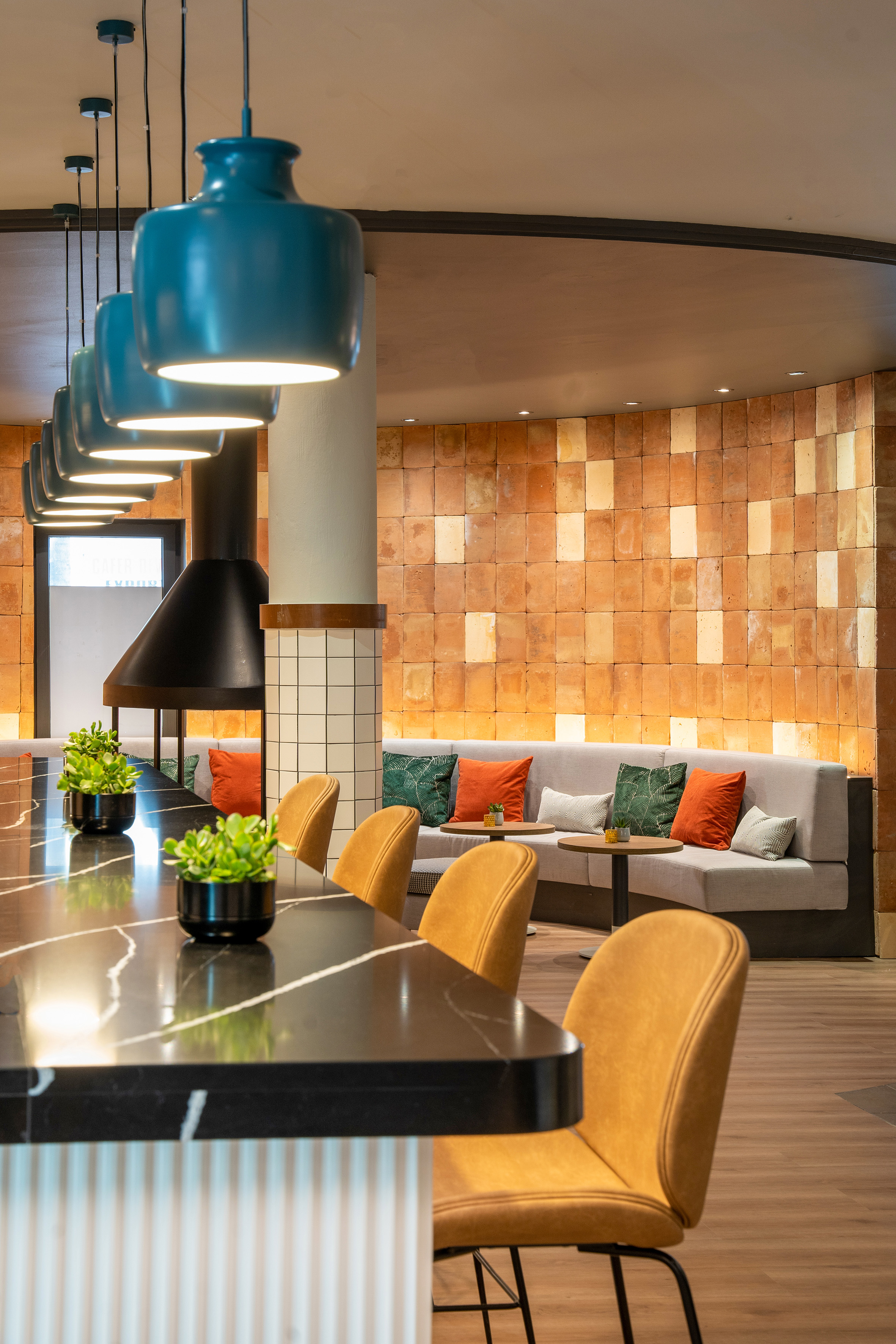 Modern bar lounge with teal pendant lights, marble counter, mustard stools, terracotta wall.