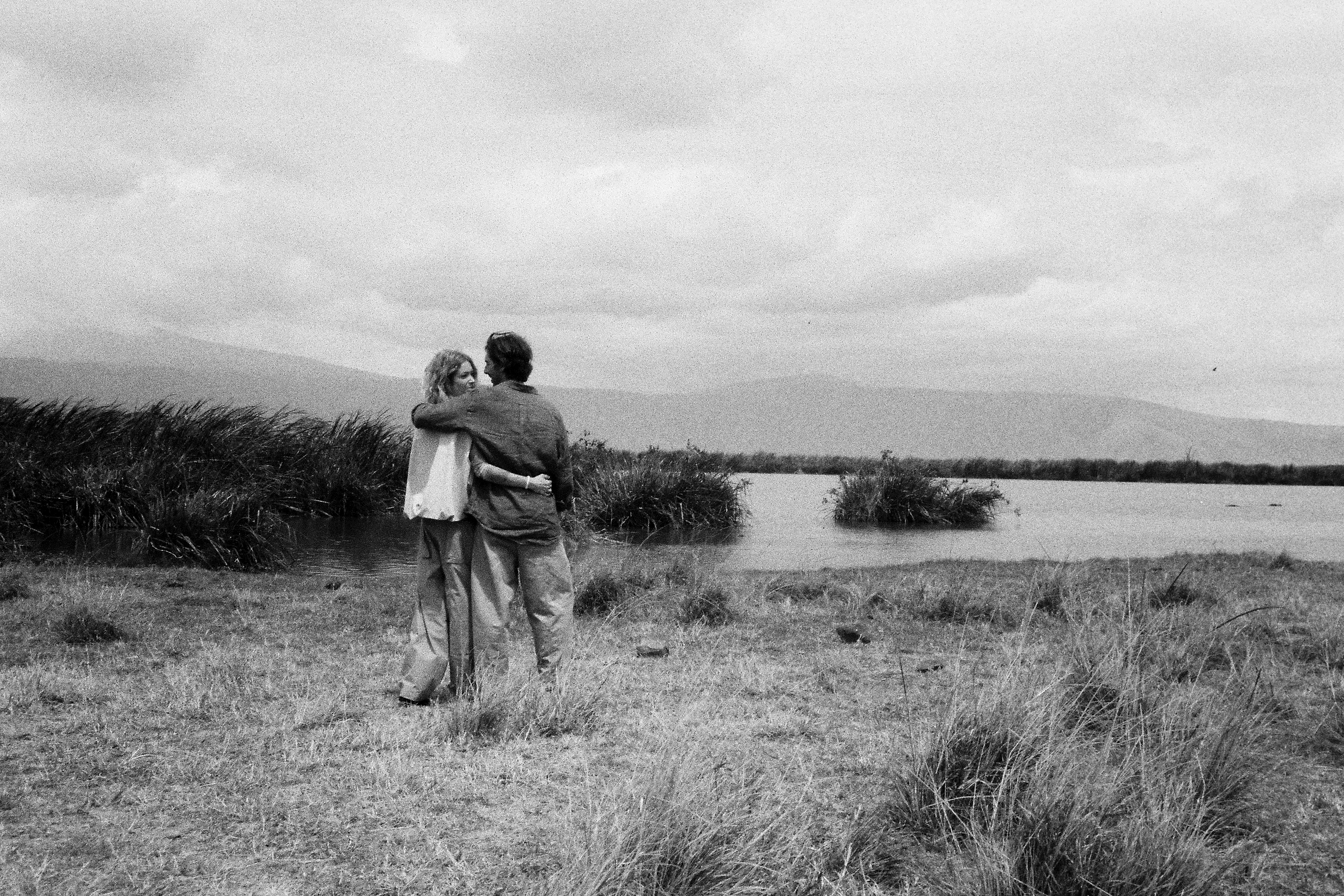 Black and white couple embracing in grassy field, lakeside with mountains.