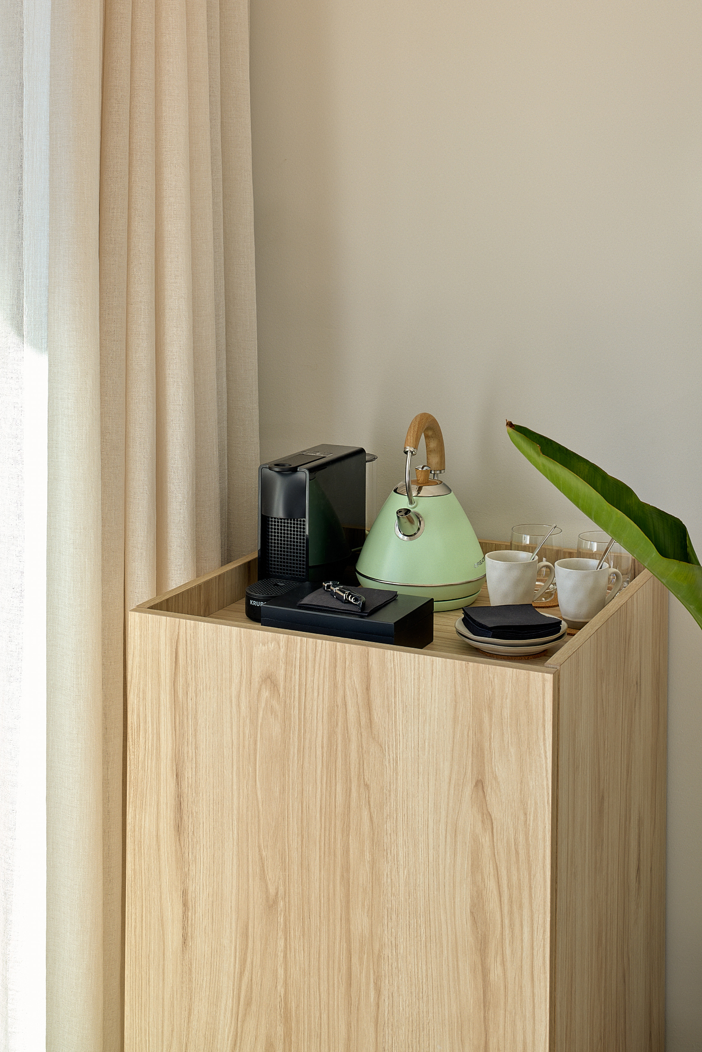 Modern coffee and tea station: green kettle, coffee machine, mugs on wood cabinet.