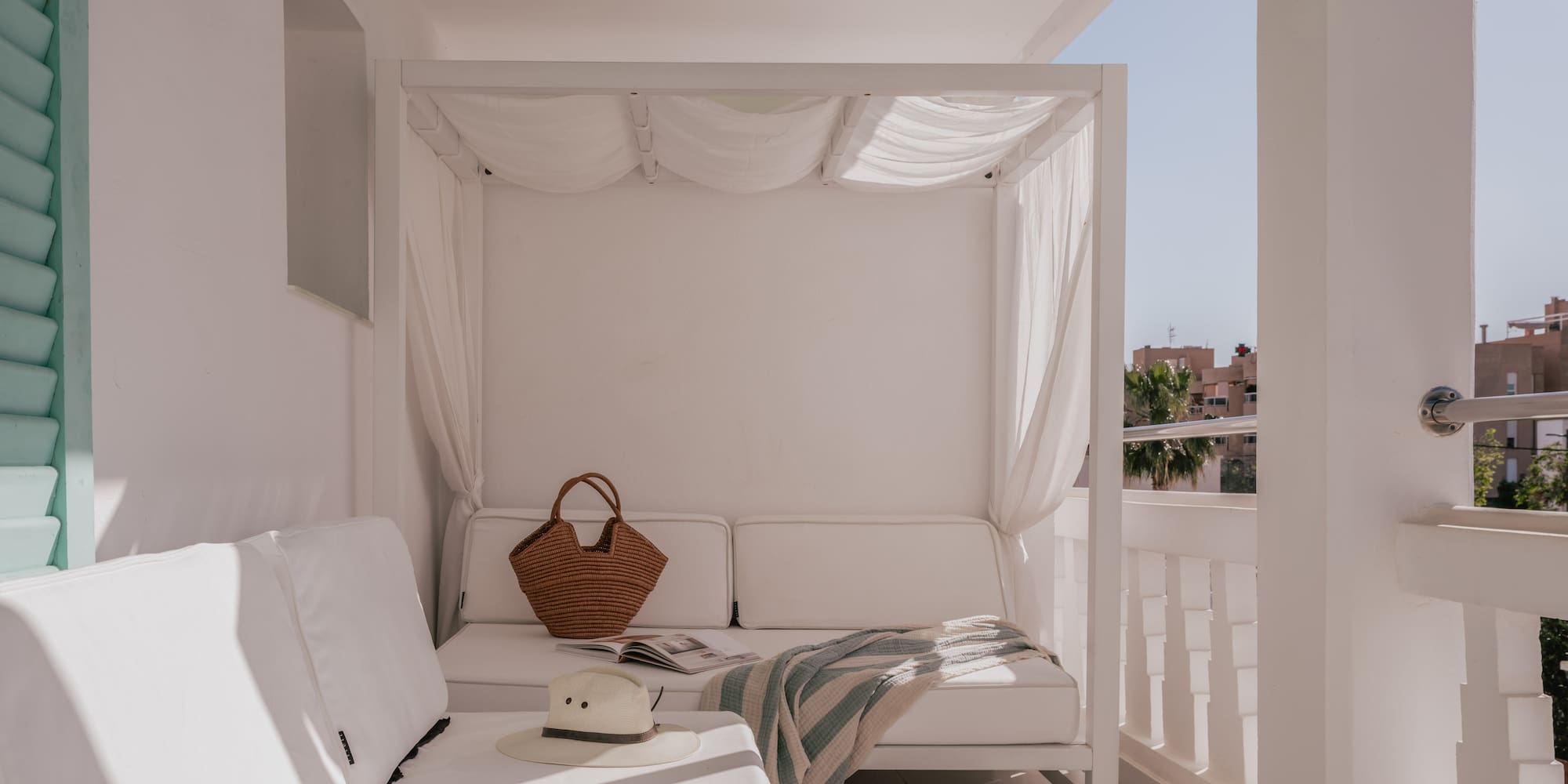 Sunny white balcony lounge with canopy daybed, straw hat, and blanket.